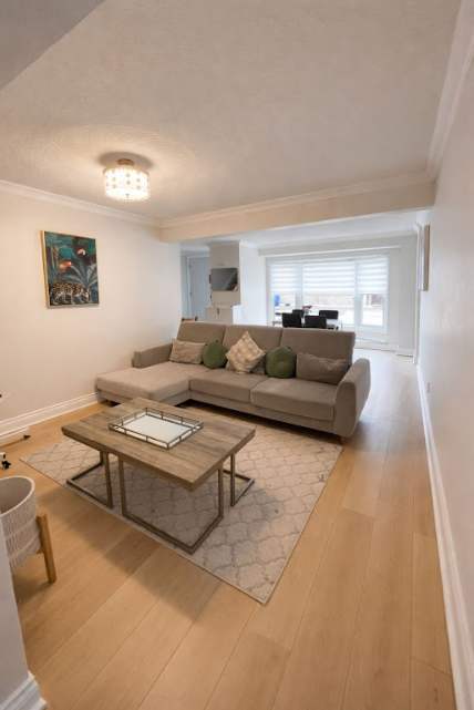 A comfortable living room featuring a large L-shaped sectional sofa and a wooden coffee table centered on a patterned area rug. The space has light wood-look flooring and an open view towards a dining area and front windows.