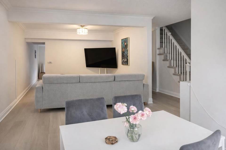 An open-concept living space with light wood-look flooring, featuring a comfortable grey sofa facing a wall-mounted television. A dining area with a white table and grey chairs is visible in the foreground, and a staircase leads to the upper level.