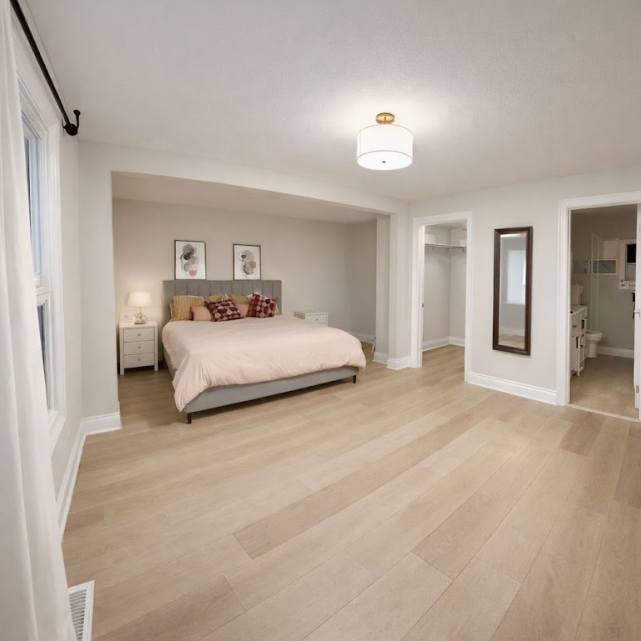 A wide-angle view of a spacious bedroom with light wood-look flooring, featuring a bed with a grey upholstered headboard, two nightstands, and access to a walk-in closet and an ensuite bathroom.