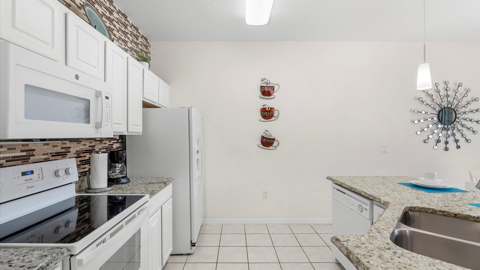 This view of the kitchen highlights the white cabinets, white appliances, and granite countertops with a mosaic tile backsplash. Decorative coffee cup wall art adorns the far wall.