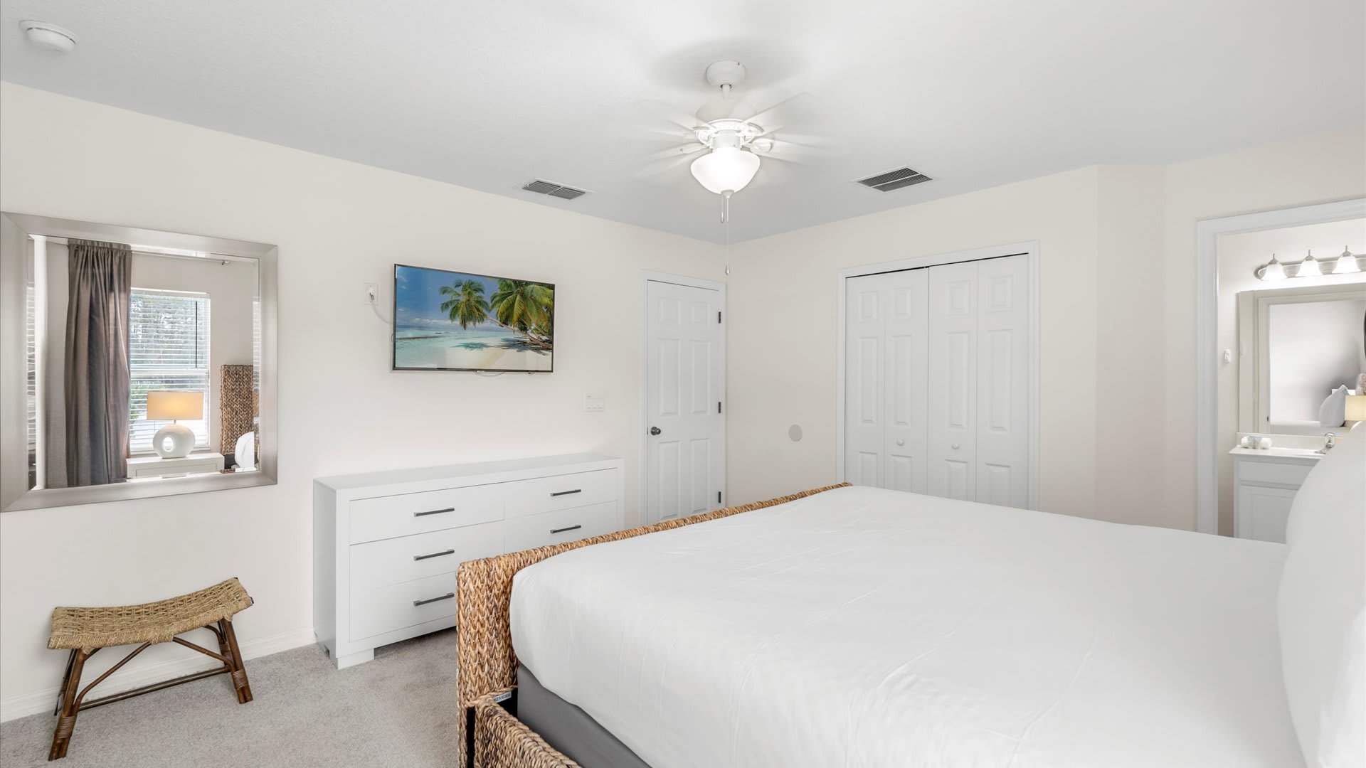 Another view of the bedroom shows a white dresser with a flat-screen TV mounted above it, a small woven stool, and a large mirror reflecting the window. A closet with bi-fold doors is also visible.
