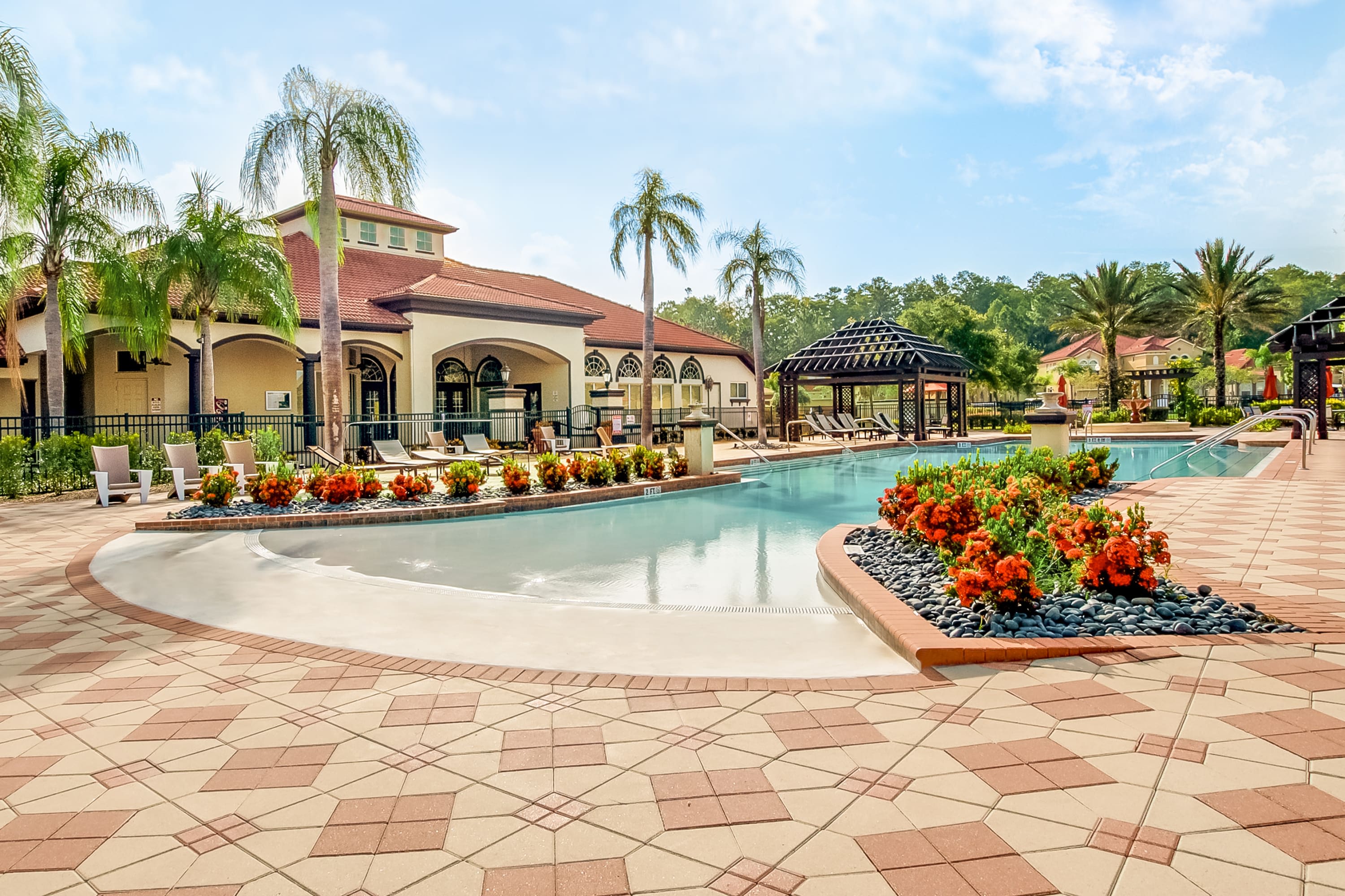 A spacious communal outdoor pool area featuring a large, irregularly shaped pool surrounded by patterned paving, lush landscaping, and a clubhouse building in the background.