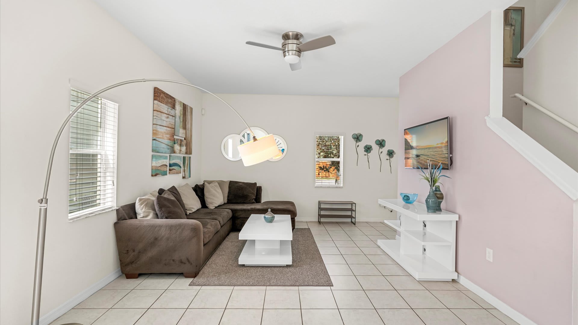 A spacious living room with a brown sectional sofa, a white coffee table, and a wall-mounted TV, featuring a light pink accent wall and stairs leading to the upper floor.