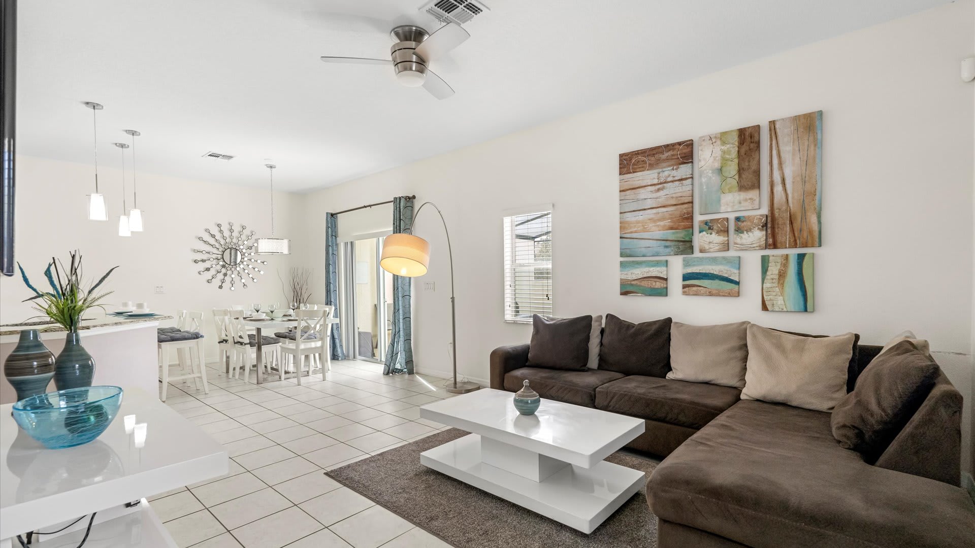 An open-concept living and dining area featuring a brown sectional sofa, a white coffee table, and a dining set, all on white tiled floors, with modern art and pendant lighting.