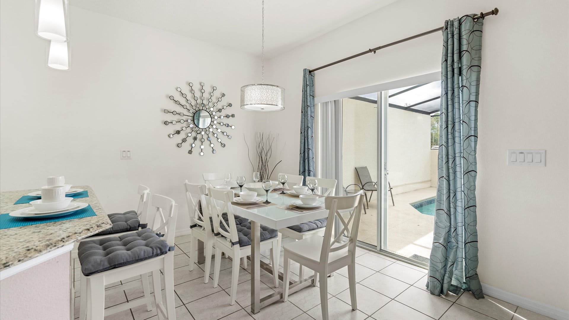 A bright dining area with a white rectangular table and six chairs, complemented by a breakfast bar with stools, and a sliding glass door opening to the outdoor pool area.