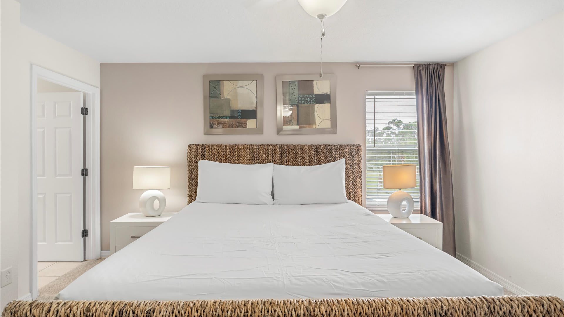 A serene bedroom features a large bed with a woven headboard and footboard, crisp white bedding, and two white nightstands with matching lamps. Two abstract art pieces hang above the bed.
