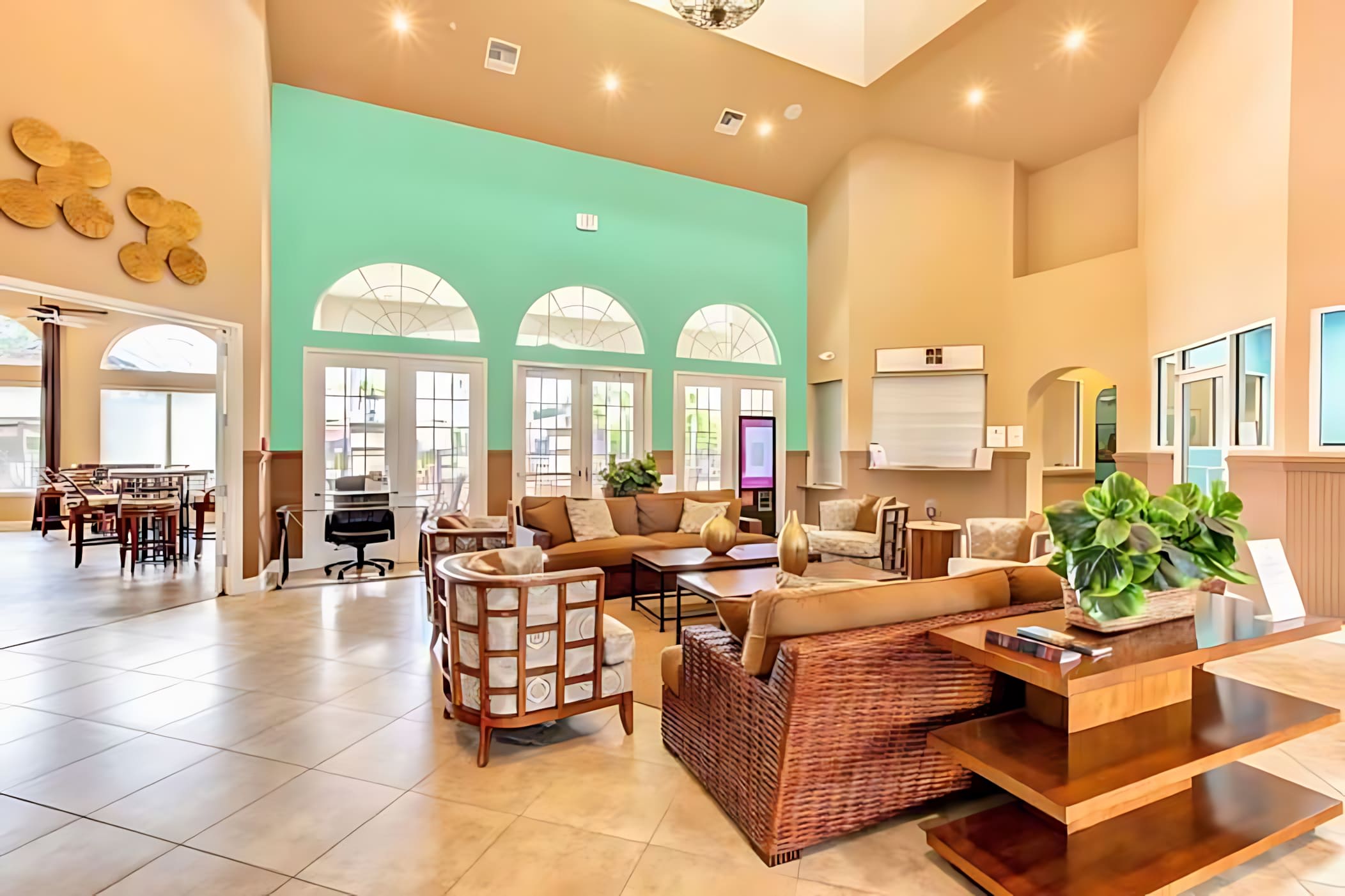 A spacious and brightly lit common area or lobby with high ceilings, featuring multiple seating arrangements with wicker and upholstered furniture, a reception desk, and large arched windows leading to an outdoor area. The walls are painted in a mix of beige and a vibrant mint green.