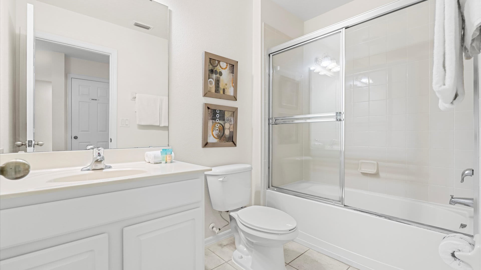 A clean bathroom with a white vanity and sink, a toilet, and a shower-tub combination enclosed by sliding glass doors, all set against white tiled walls and floor.