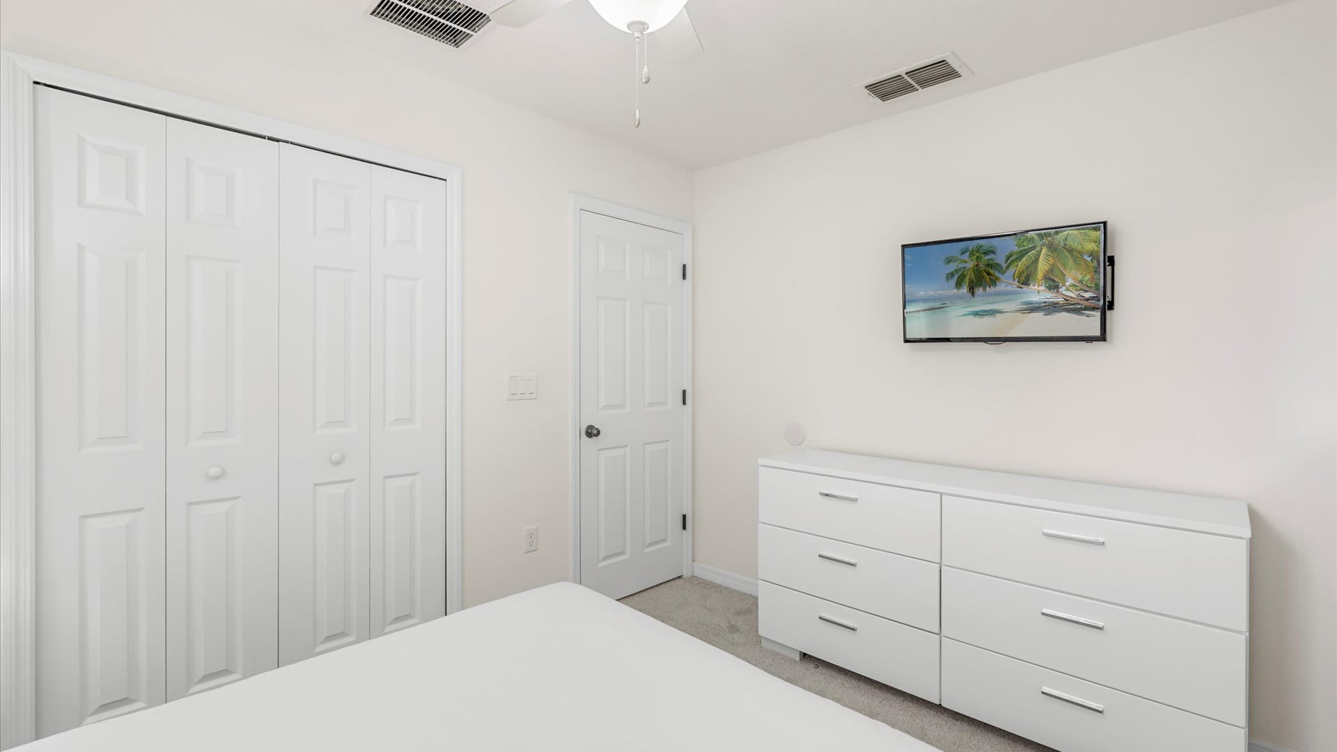 This bedroom view shows a white dresser with a flat-screen TV mounted above it, a closet with white bi-fold doors, and a white door leading to another room. The floor is carpeted.
