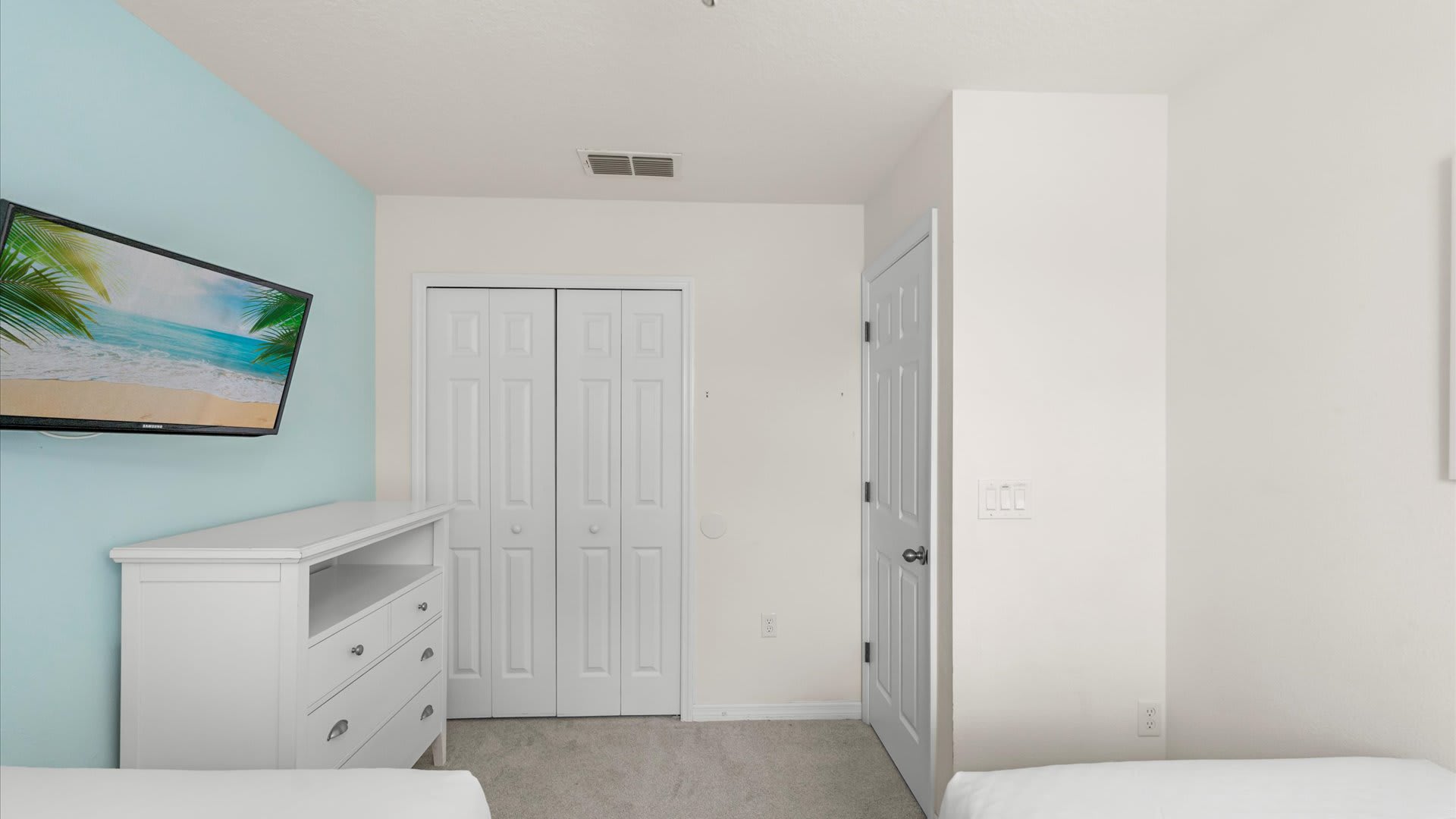 A corner view of the twin bedroom, showing a wall-mounted TV on a light blue accent wall, a white dresser below it, and a white bi-fold closet door.