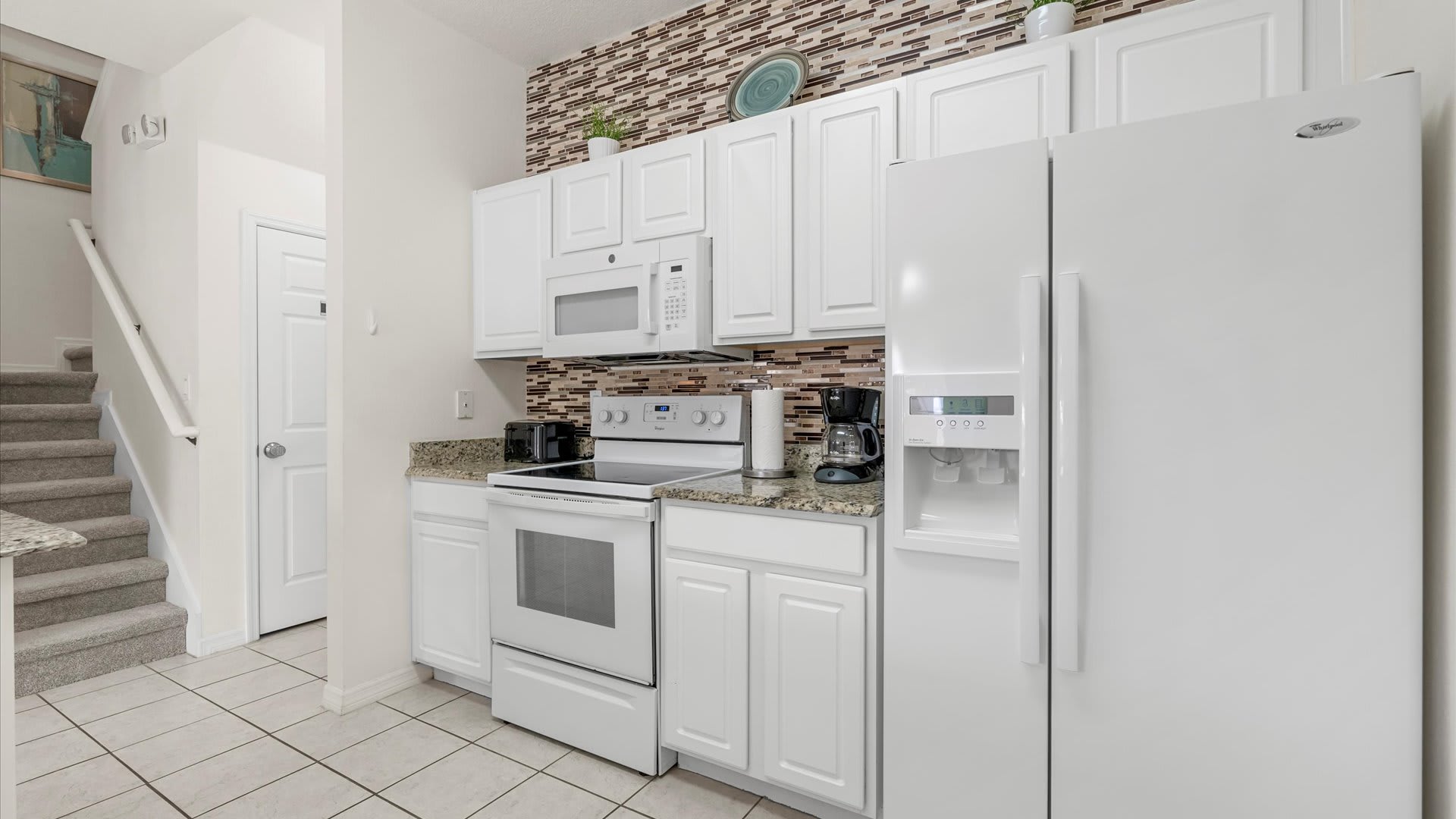 A bright kitchen features white cabinetry, white appliances, granite countertops, and a brown and white mosaic tile backsplash. A carpeted staircase is visible on the left.