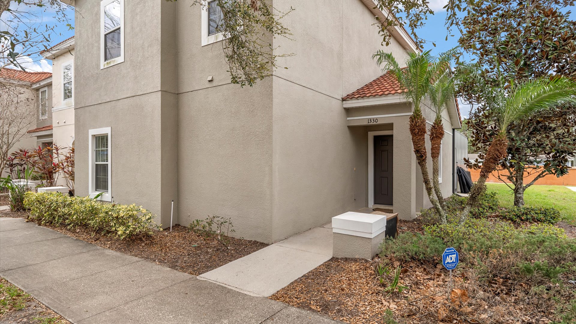 An angled view of the townhouse exterior, highlighting the light beige stucco walls, red tile roof, and the front entrance with the number 1330, surrounded by green landscaping.