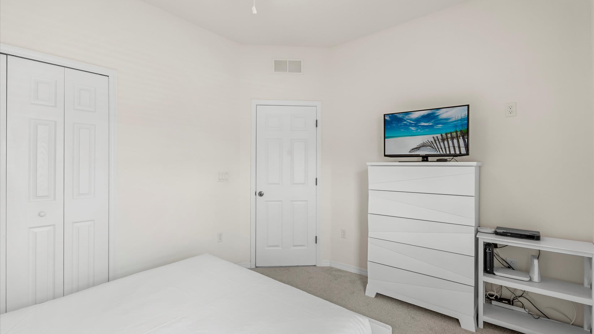A bright bedroom with white walls and carpet, featuring a white dresser with a TV displaying a beach scene, and a white bed with a simple white duvet.