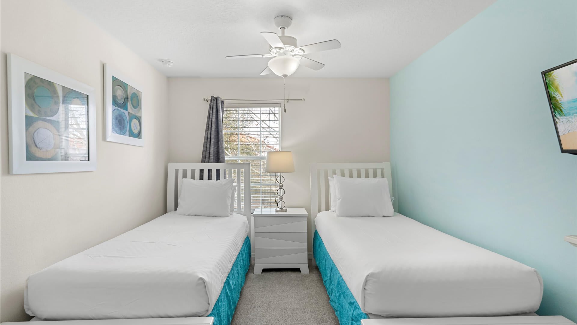 A twin bedroom featuring two white-framed beds with blue bed skirts, a light blue accent wall, a ceiling fan, and a white nightstand with a lamp between the beds.