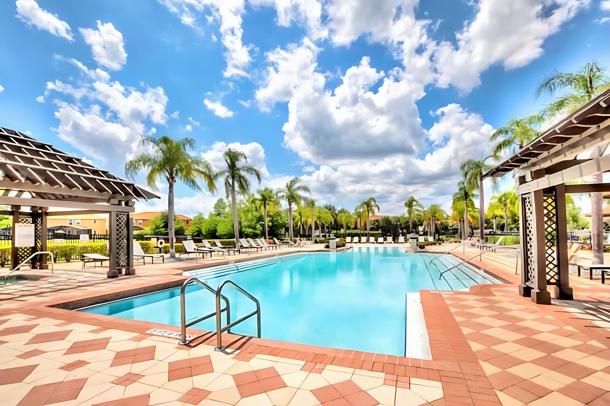 A large, bright blue communal swimming pool surrounded by a patterned red and beige tiled deck, with multiple lounge chairs, palm trees, and two wooden pergolas under a partly cloudy sky.