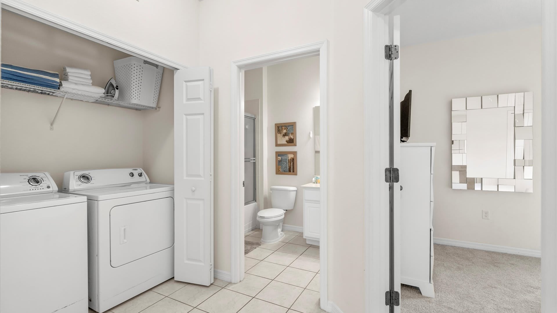 A functional laundry area with a white washer and dryer, shelving with towels, and an open doorway leading into a bathroom with a toilet and shower.