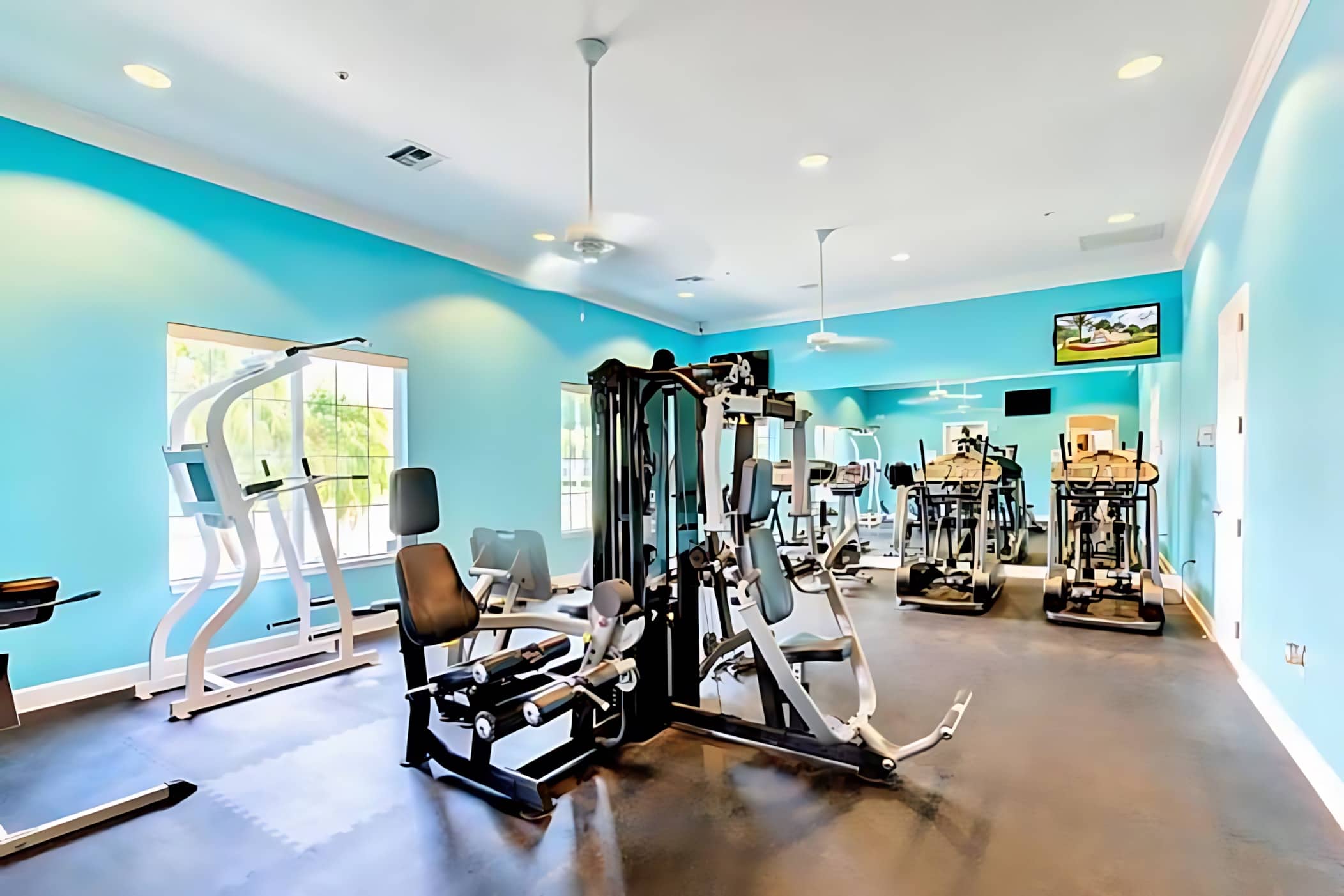 A bright fitness center with light blue walls, featuring multiple exercise machines including treadmills, weight machines, and elliptical trainers, with large mirrors and ceiling fans.