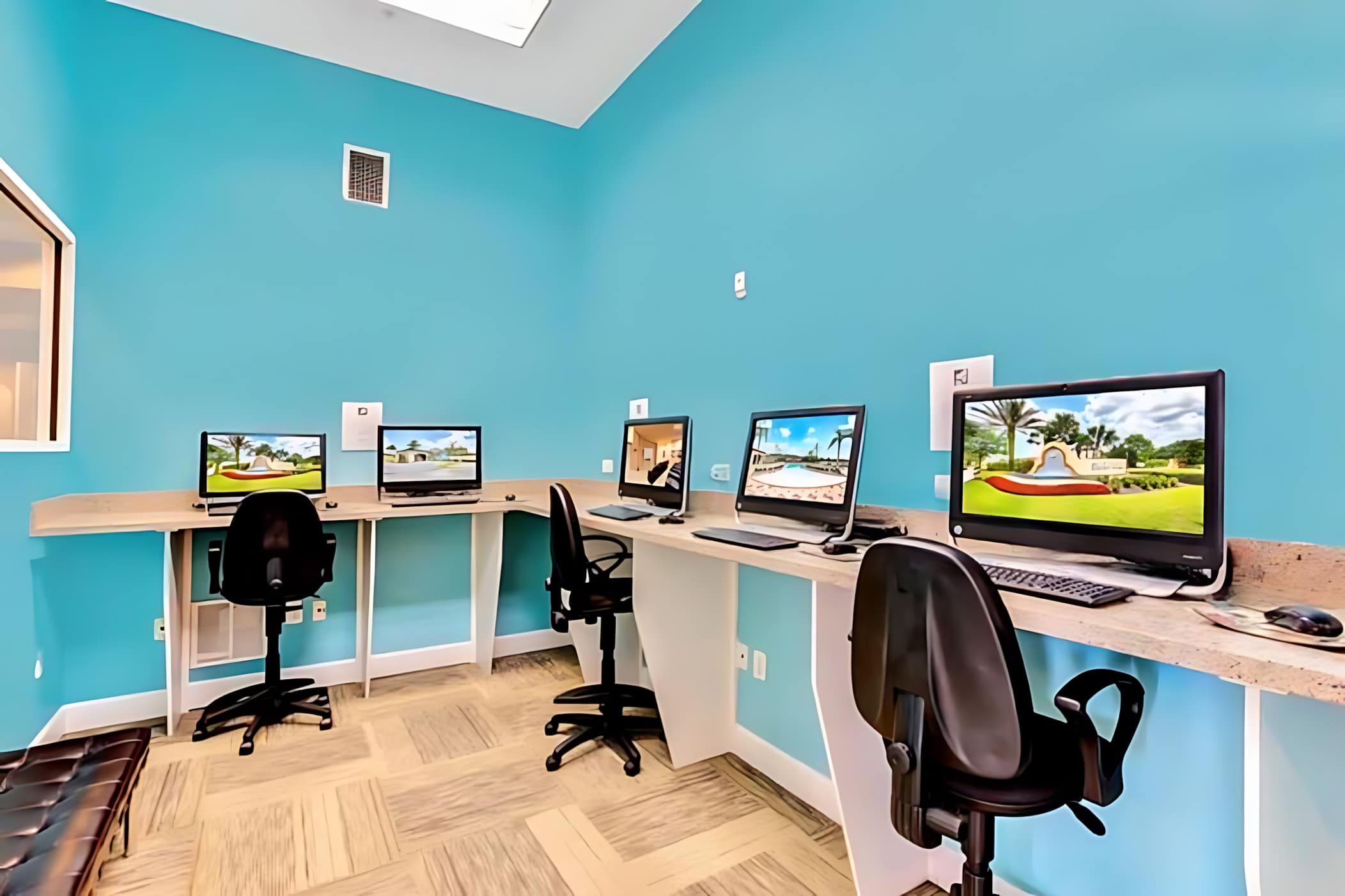 A modern business center with light blue walls, featuring several computer workstations with desktop computers, office chairs, and a long light-colored counter, set on a patterned carpeted floor.