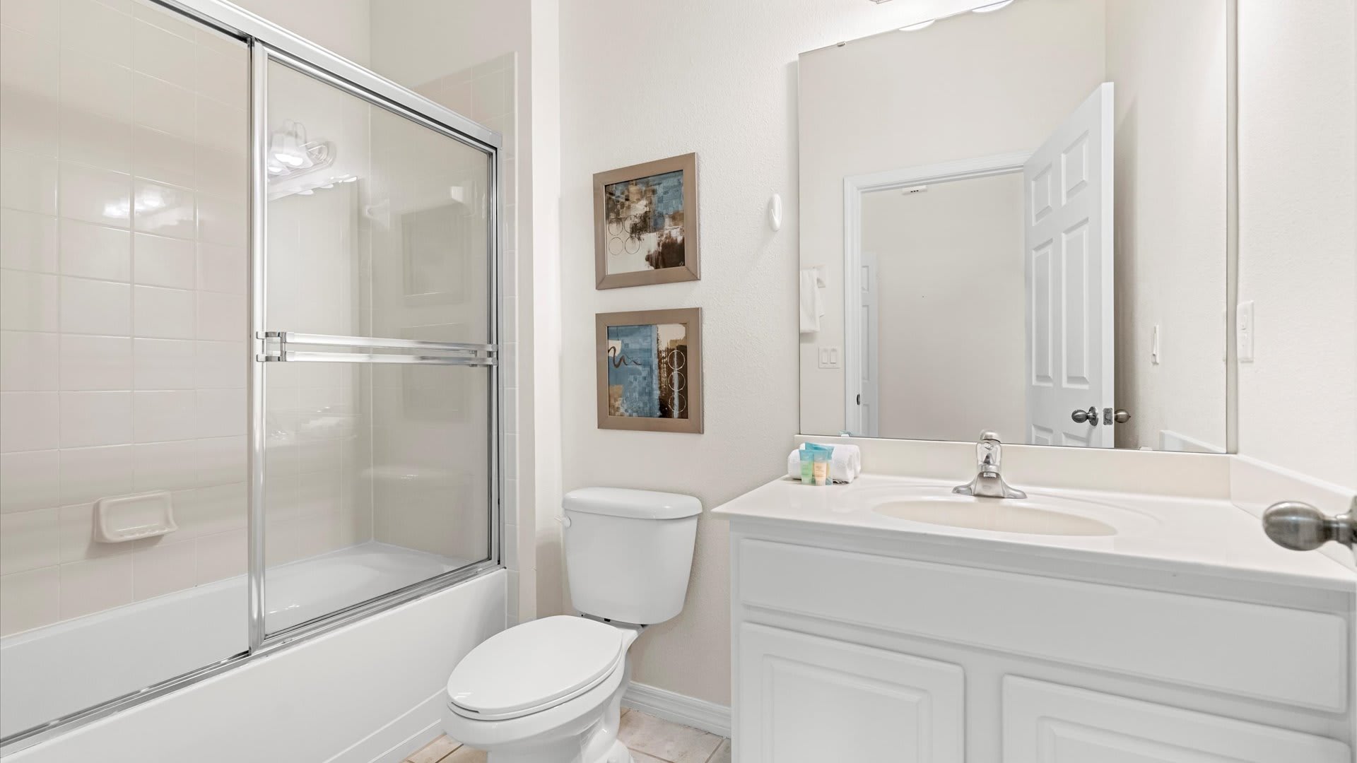 A clean bathroom with a white vanity, single sink, and a large mirror. It includes a toilet and a bathtub with a sliding glass shower door, set against light tiled walls.