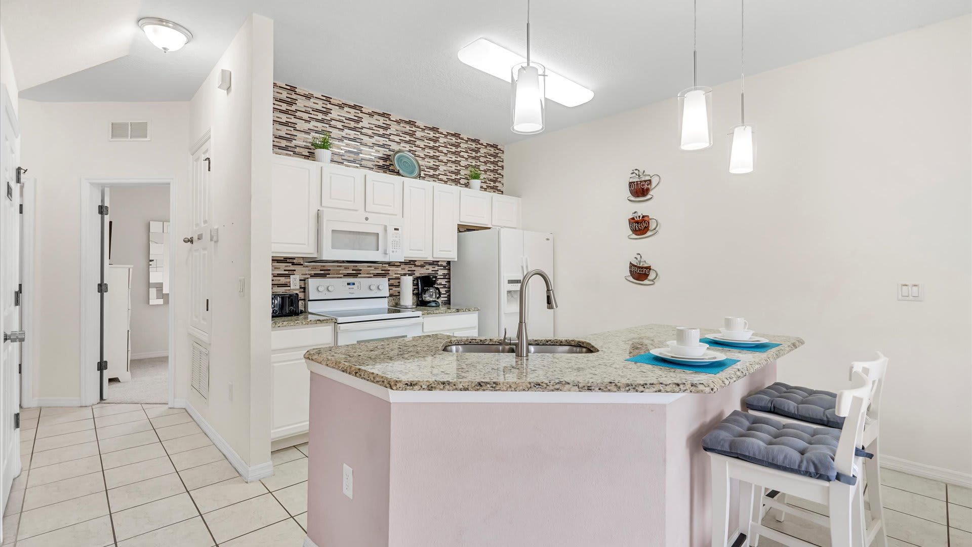 A spacious kitchen with white cabinets, white appliances, and granite countertops, featuring a breakfast bar with two stools. A stainless steel sink and modern faucet are integrated into the island.