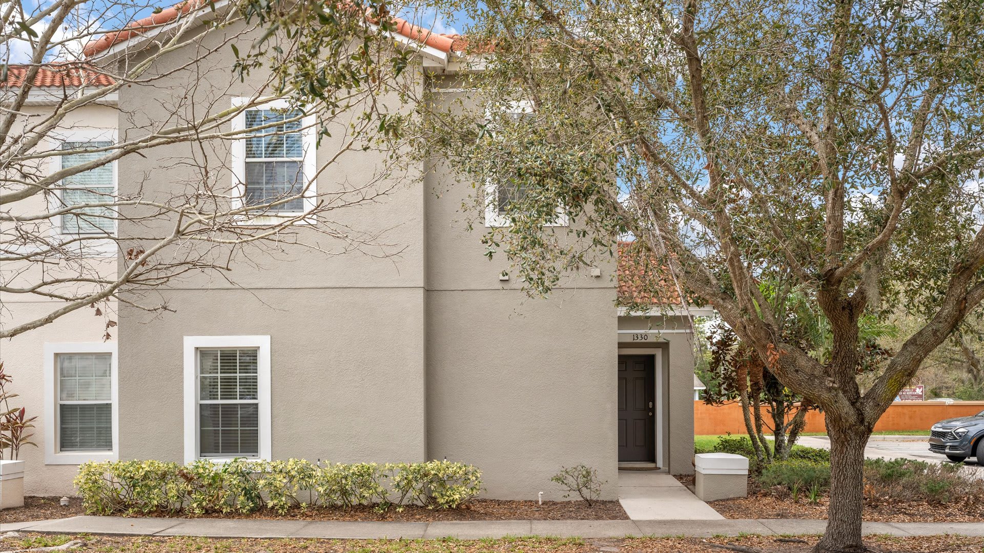 The front exterior of a two-story townhouse with a light beige stucco facade, a dark brown front door, and a red tile roof, surrounded by mature trees and landscaping.