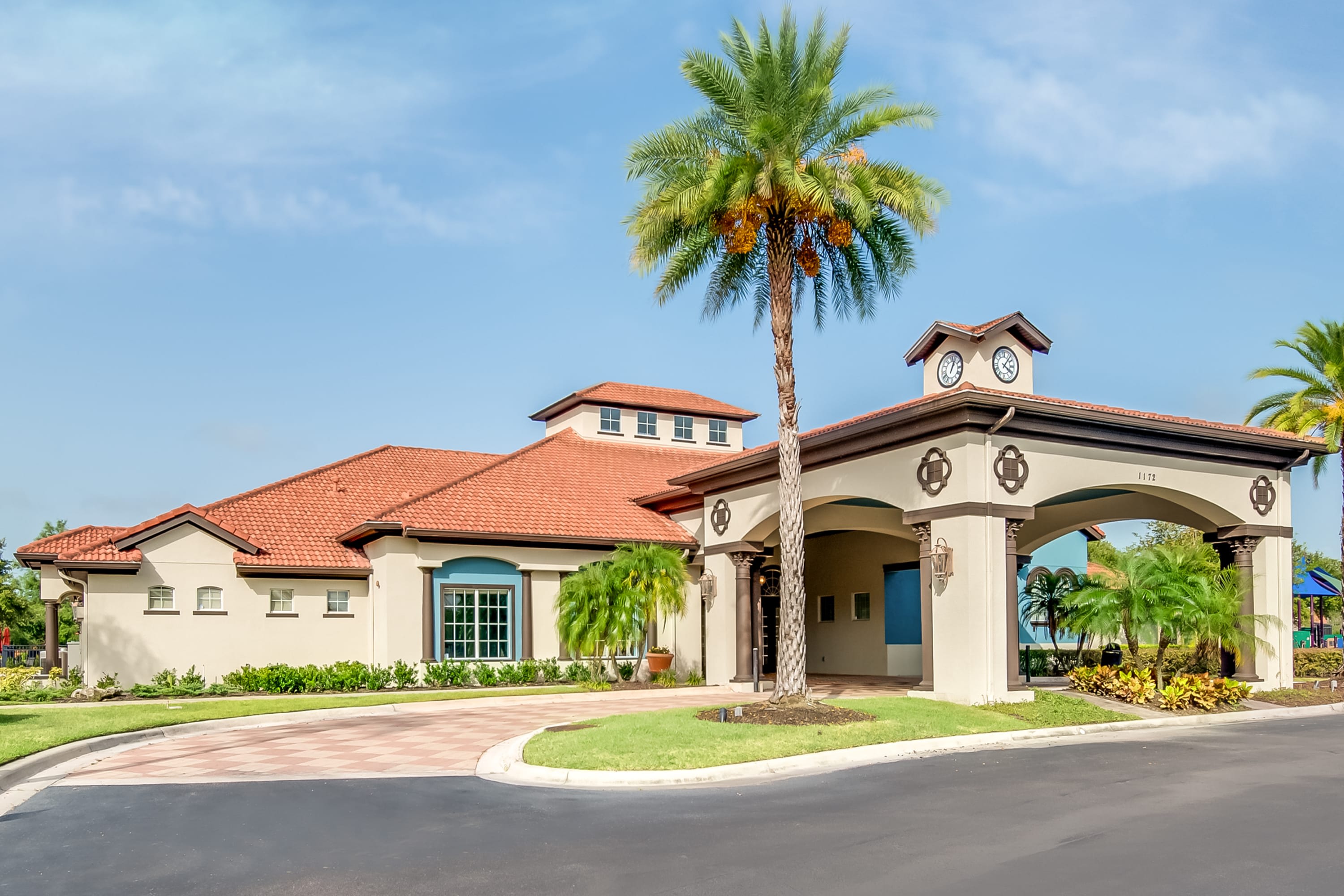 The grand entrance to a resort clubhouse, featuring a large archway with a clock tower, a paved driveway, and lush landscaping with palm trees under a clear blue sky.