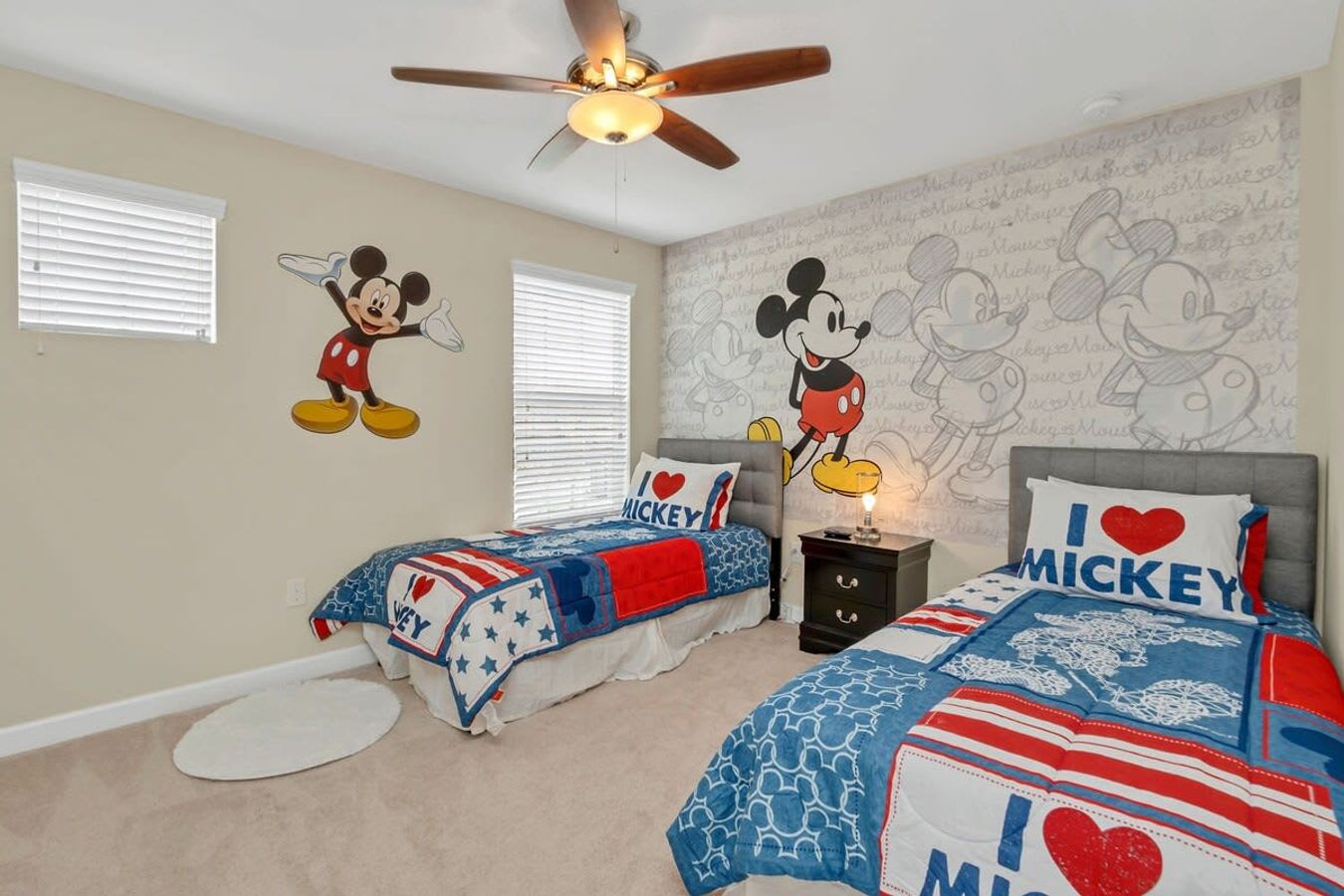 A vibrant Mickey Mouse-themed bedroom with two twin beds featuring red, white, and blue bedding, set against light beige walls with large Mickey Mouse decals and a patterned accent wall. A ceiling fan with a light fixture is centered above.