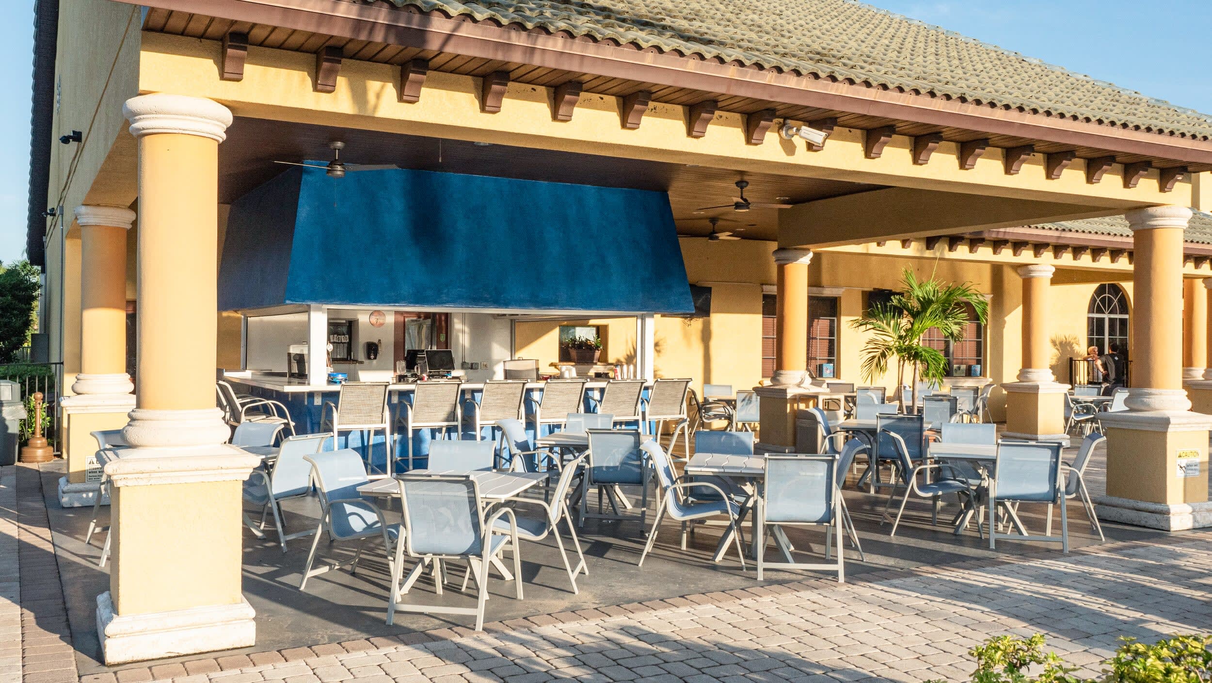 An inviting outdoor covered bar and dining area featuring a blue bar counter with stools, surrounded by numerous light blue tables and chairs on a paved patio, supported by yellow columns.