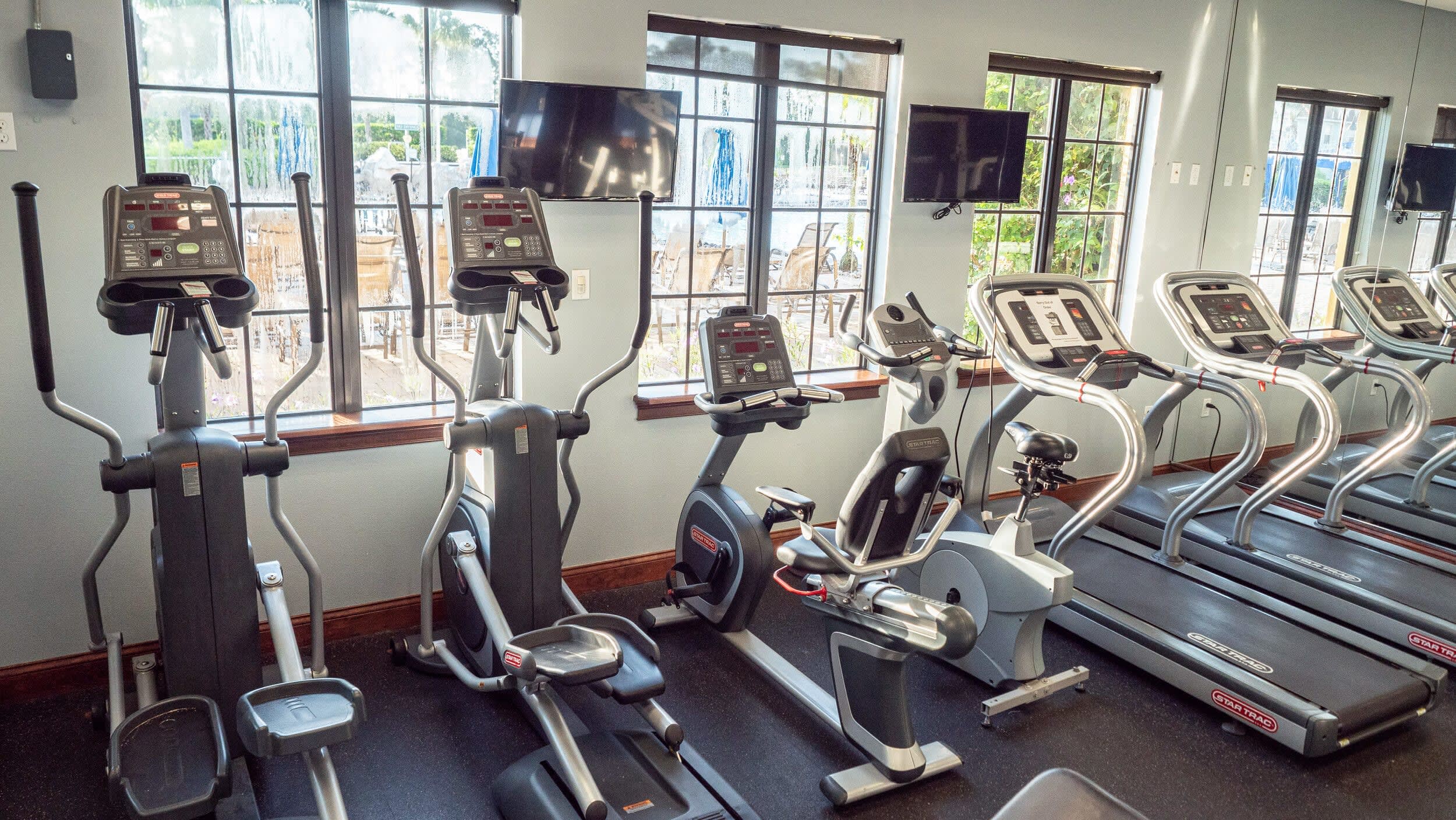 An indoor fitness center equipped with several modern treadmills, elliptical machines, and stationary bikes, all positioned to face large windows offering a view of an outdoor area.