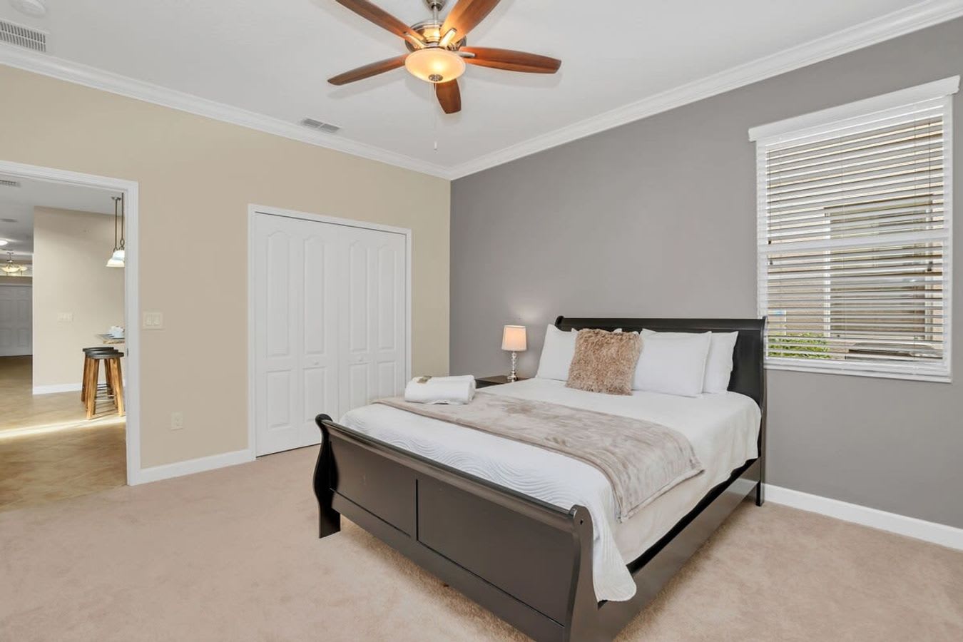 A cozy bedroom with a dark wood sleigh bed dressed in white and beige bedding, positioned against a gray accent wall. A ceiling fan with a light fixture is overhead, and a small nightstand with a lamp sits beside the bed.