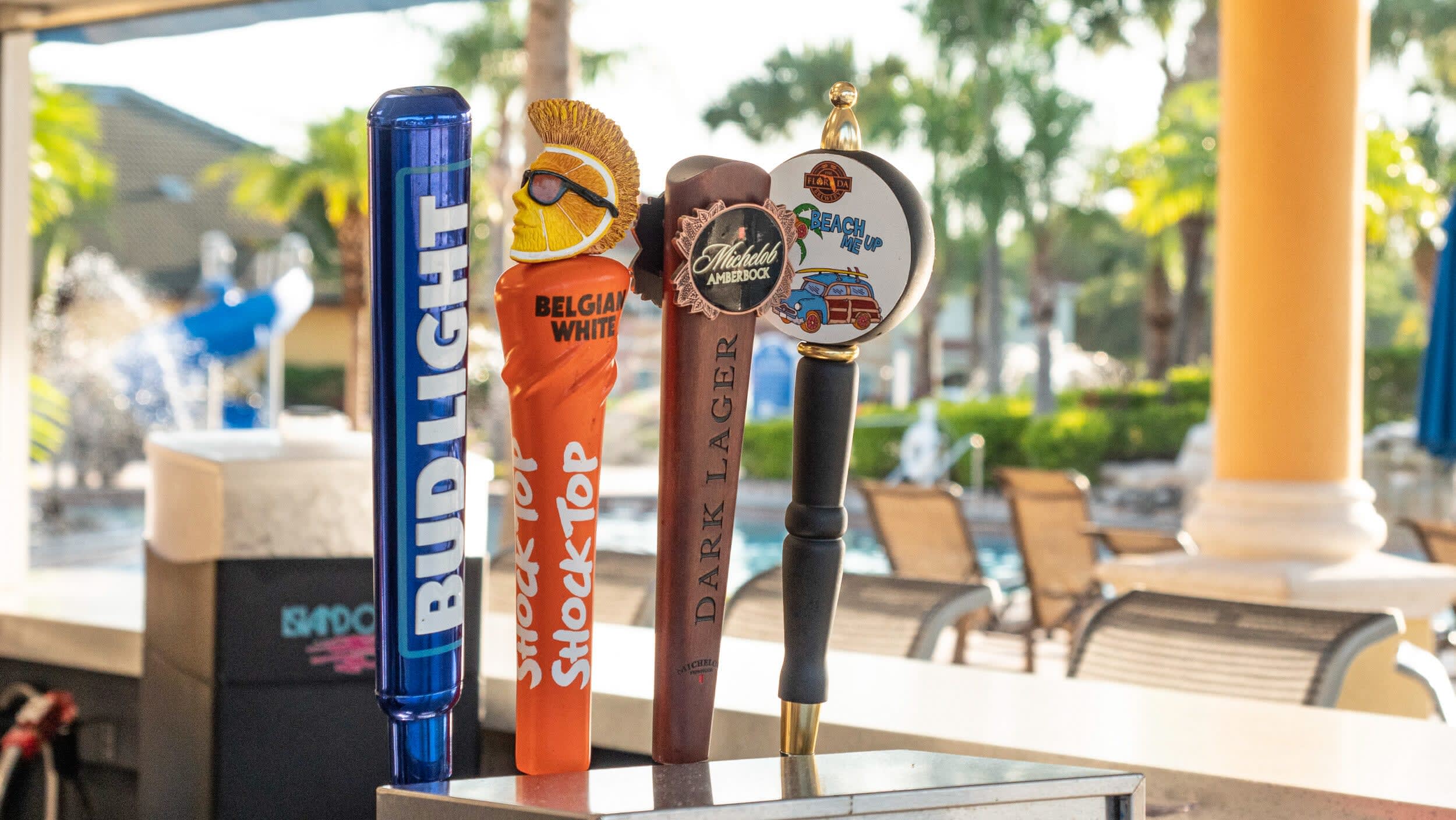 A close-up view of four distinct beer taps on a bar counter, including 'Bud Light' and 'Shock Top,' with a blurred background showing a sunny outdoor pool area and palm trees.