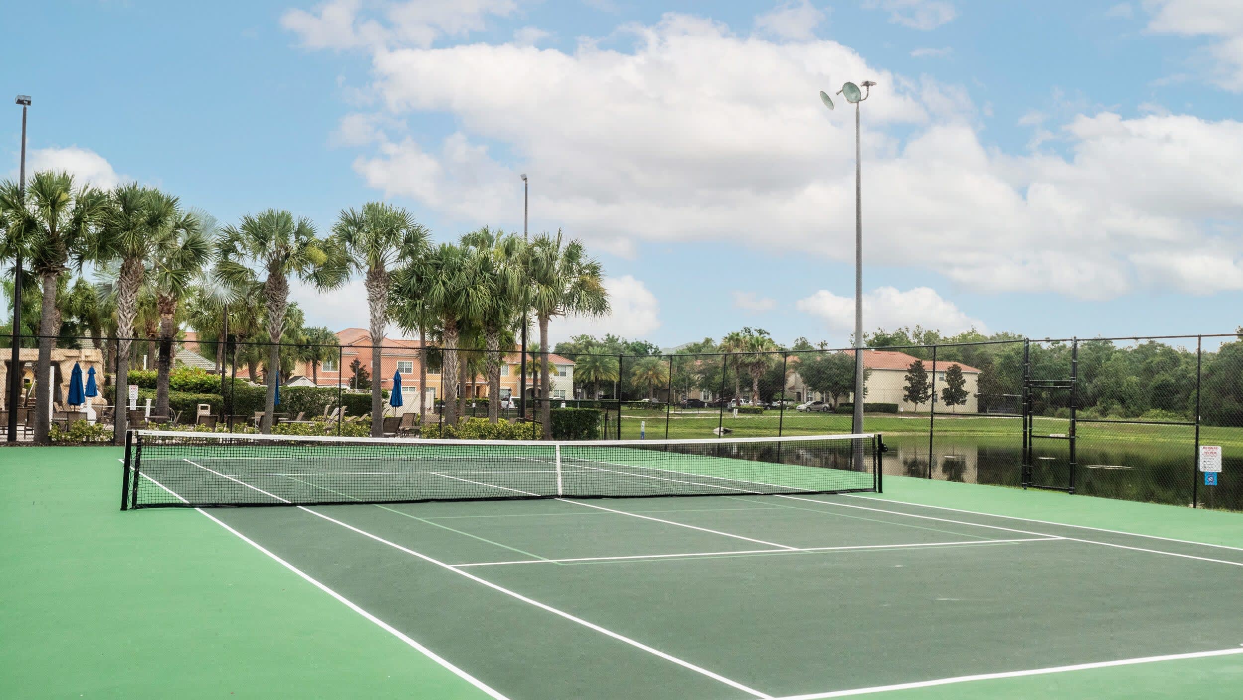An outdoor tennis court with a green surface and white lines, featuring a net in the center and surrounded by a black fence. Palm trees and resort buildings are visible in the background, with a lake beyond the fence.