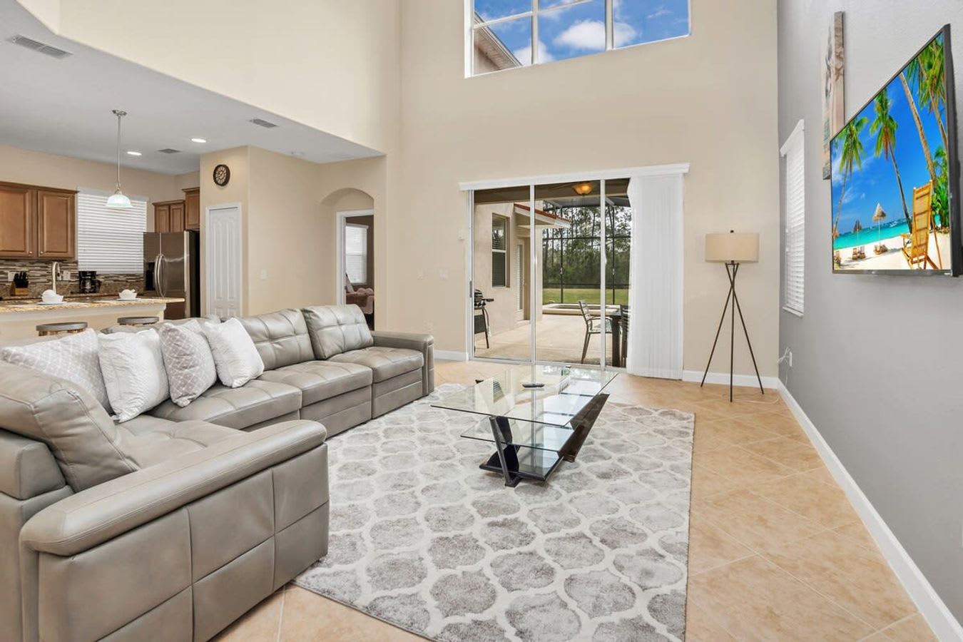 Spacious living room with a large gray sectional sofa, a glass coffee table on a patterned area rug, a wall-mounted TV, and sliding glass doors opening to a screened patio.