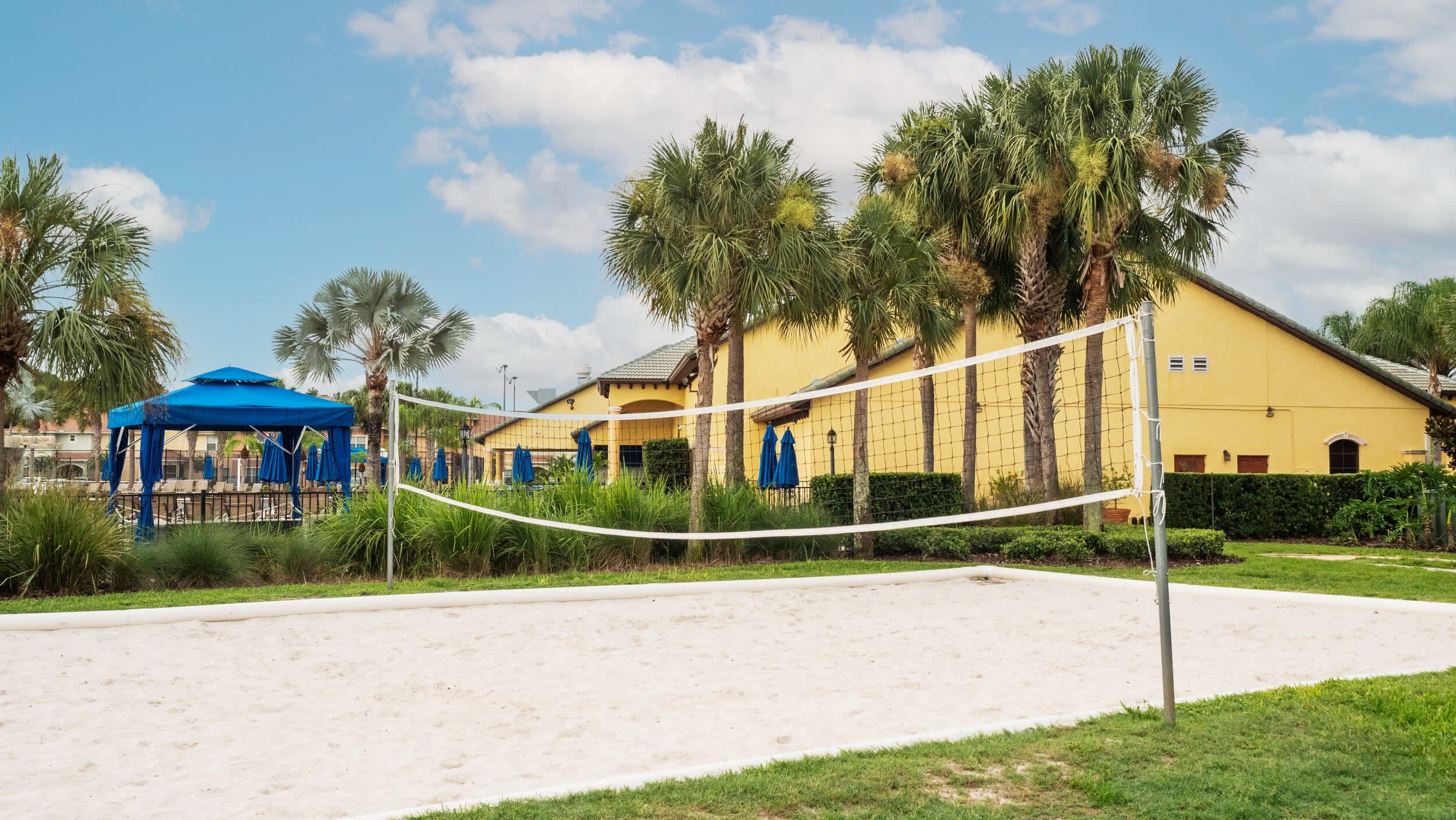 A bright outdoor sand volleyball court with a net, surrounded by green grass and tall palm trees, with a yellow building and blue cabanas in the background under a partly cloudy sky.