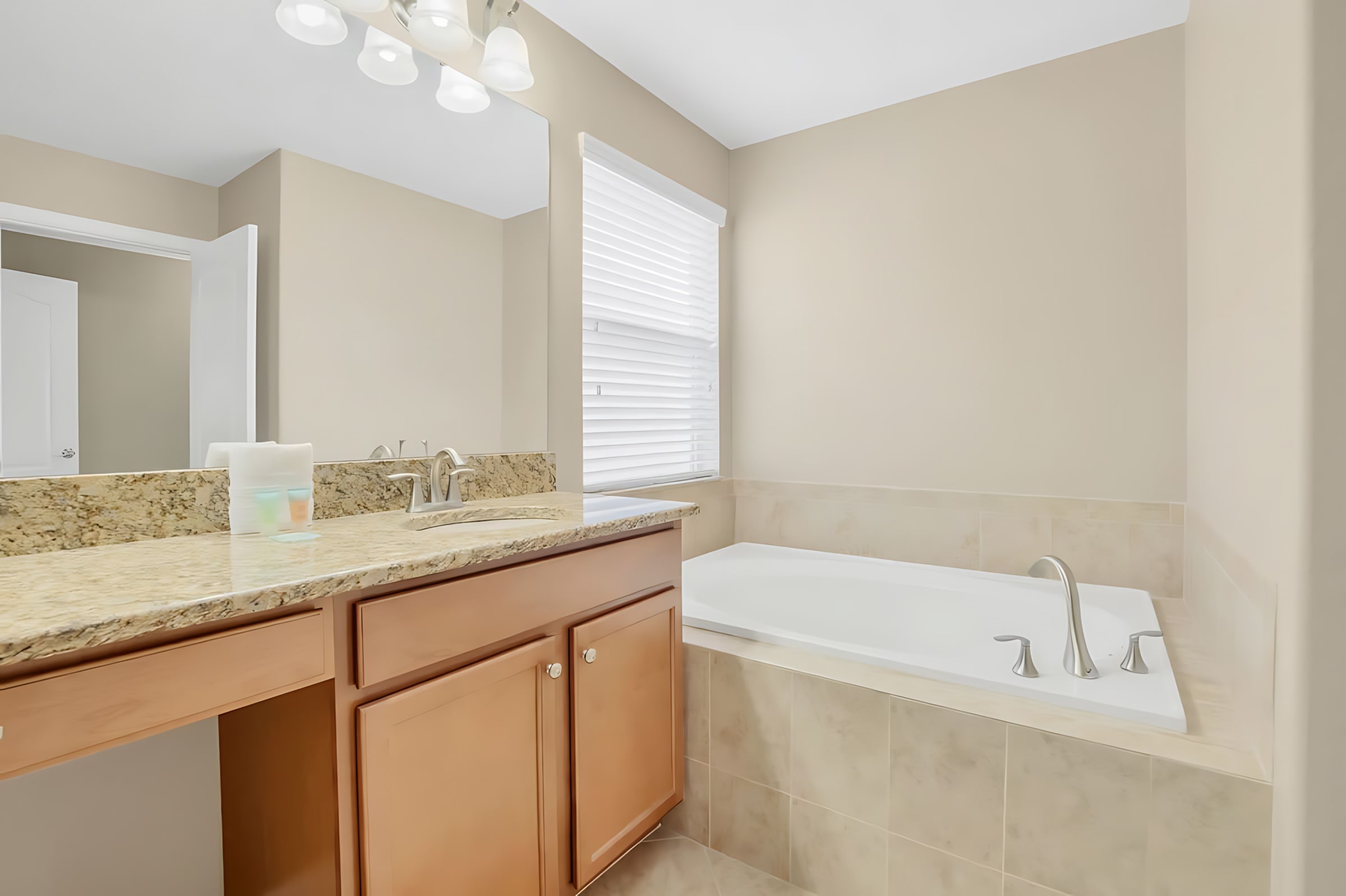 A spacious bathroom with light beige walls and tiled floors, featuring a large built-in bathtub with a tiled surround and a double vanity with a granite countertop and light wood cabinets. A large mirror and bright vanity lighting are above the sinks.