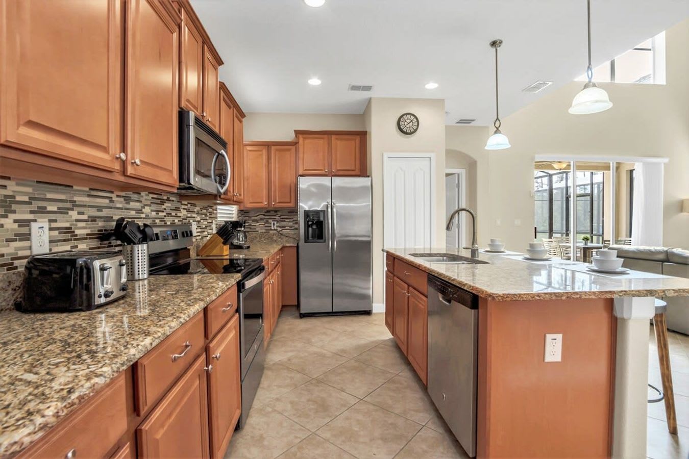 A wide view of the kitchen showcasing light brown wooden cabinetry, granite countertops, and stainless steel appliances including a double-door refrigerator and dishwasher. The kitchen island with a sink and pendant lights opens up to a view of the living area and outdoor space.