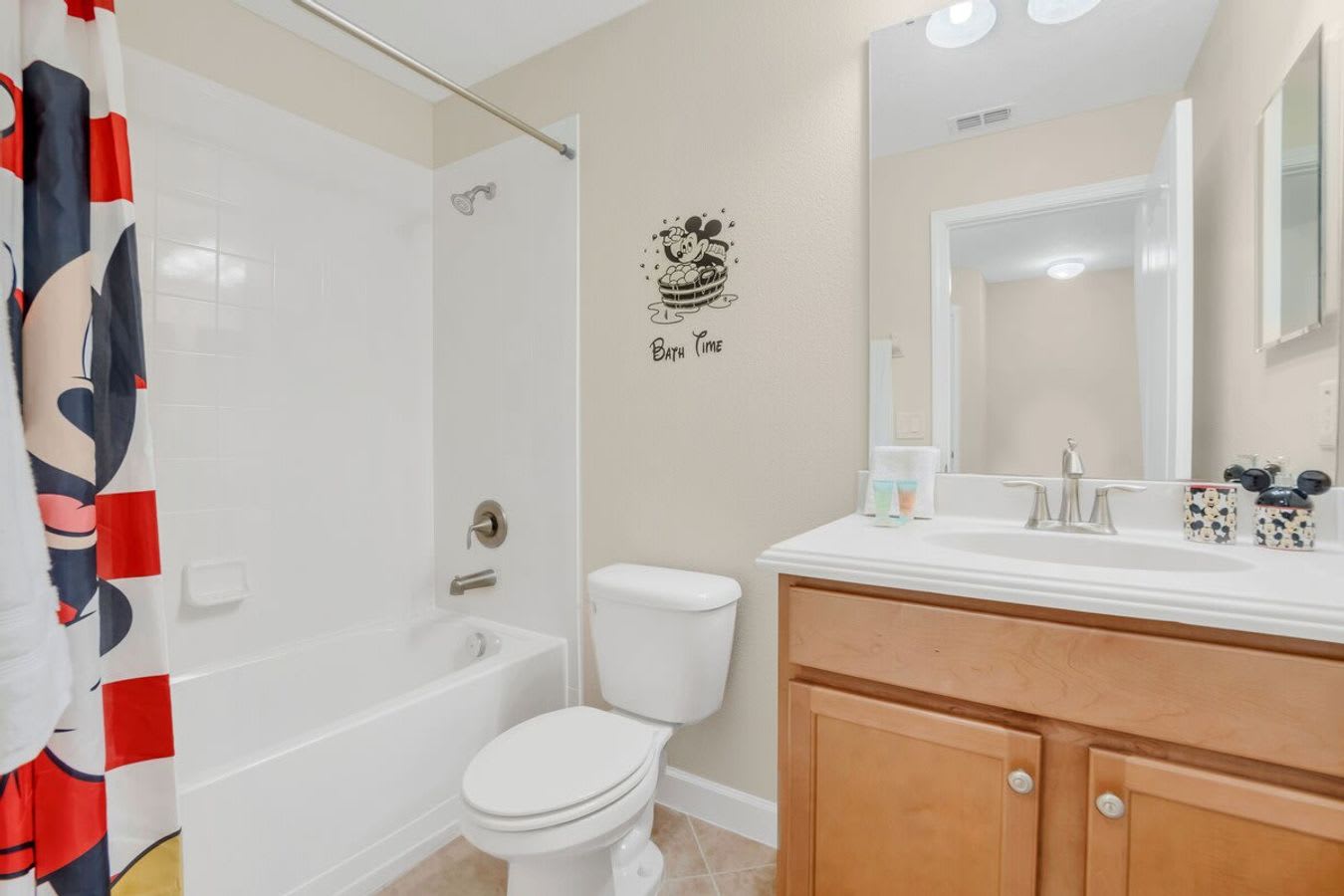 A bright bathroom with light beige walls and tiled floors, featuring a white shower/tub combo with a Mickey Mouse-themed shower curtain and a single vanity with a white countertop and light wood cabinets. A mirror and vanity lighting are above the sink.