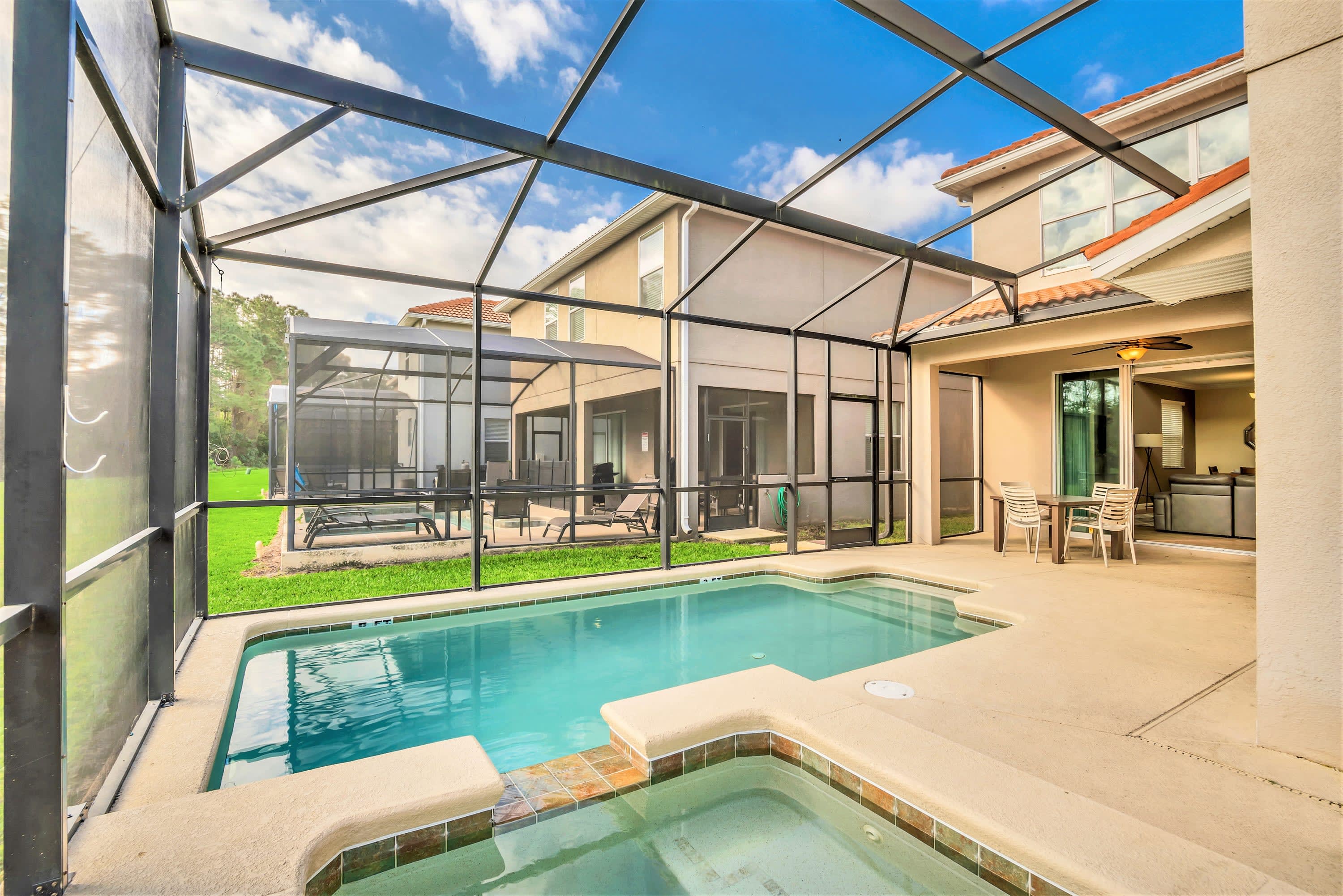 Rear view of the house showcasing a large screened-in private pool and hot tub area, with additional lounge seating, overlooking a grassy backyard and distant trees.