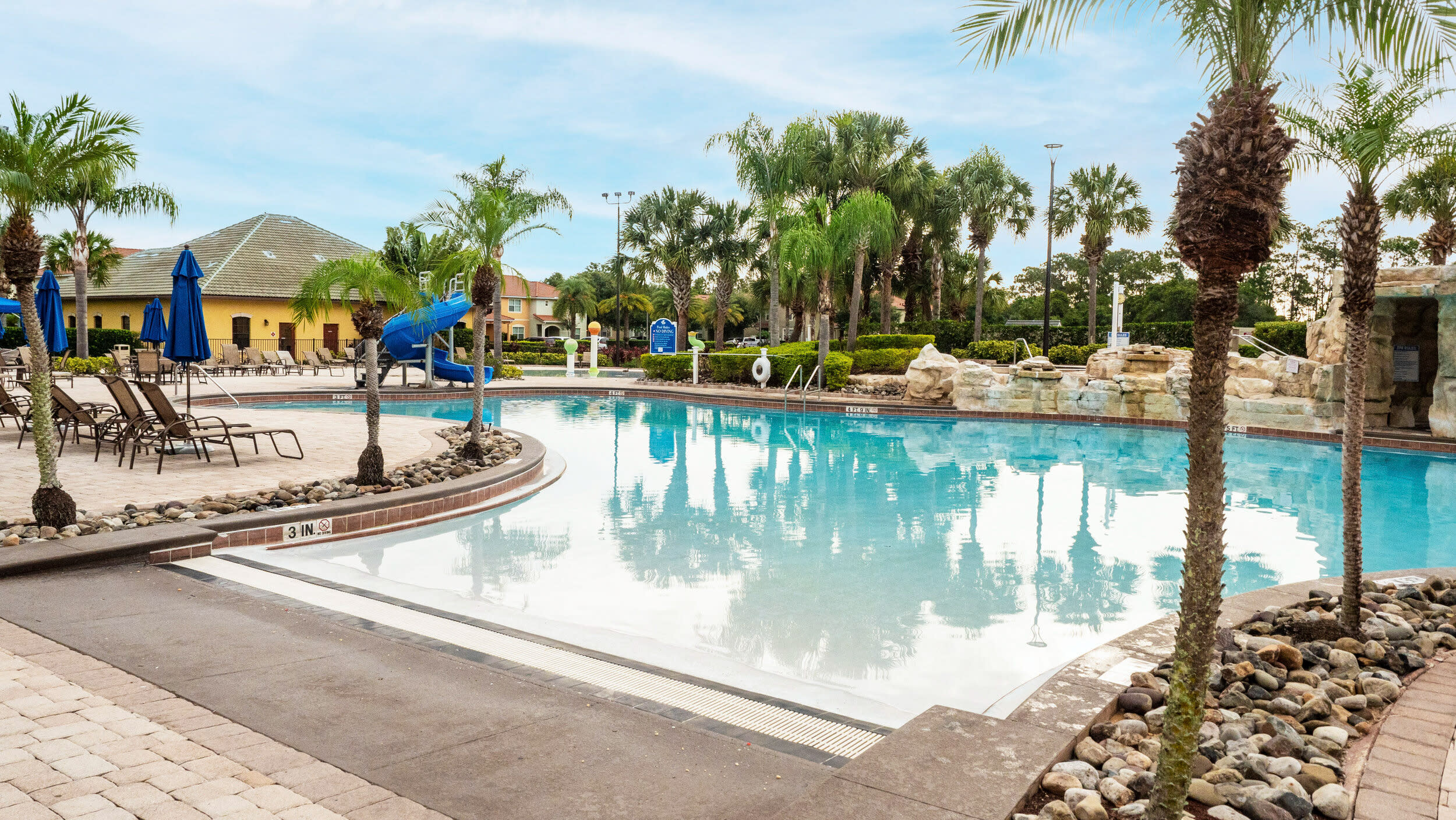 Expansive communal swimming pool area surrounded by palm trees, lounge chairs, and a decorative rock waterfall feature, under a bright blue sky.