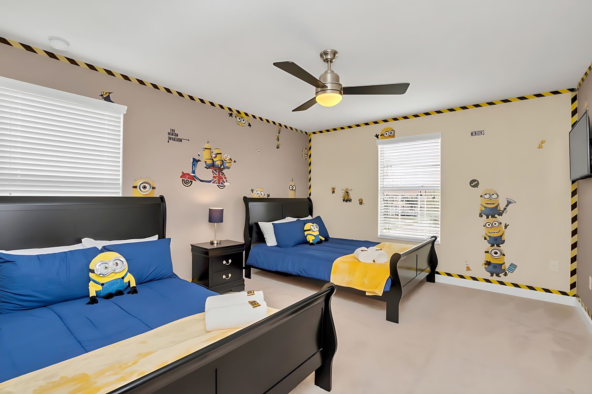 A fun Minion-themed bedroom featuring two black twin beds with blue bedding and yellow Minion pillows, set against light brown walls adorned with Minion decals and a black and yellow striped border. A ceiling fan with a light fixture is centered above.