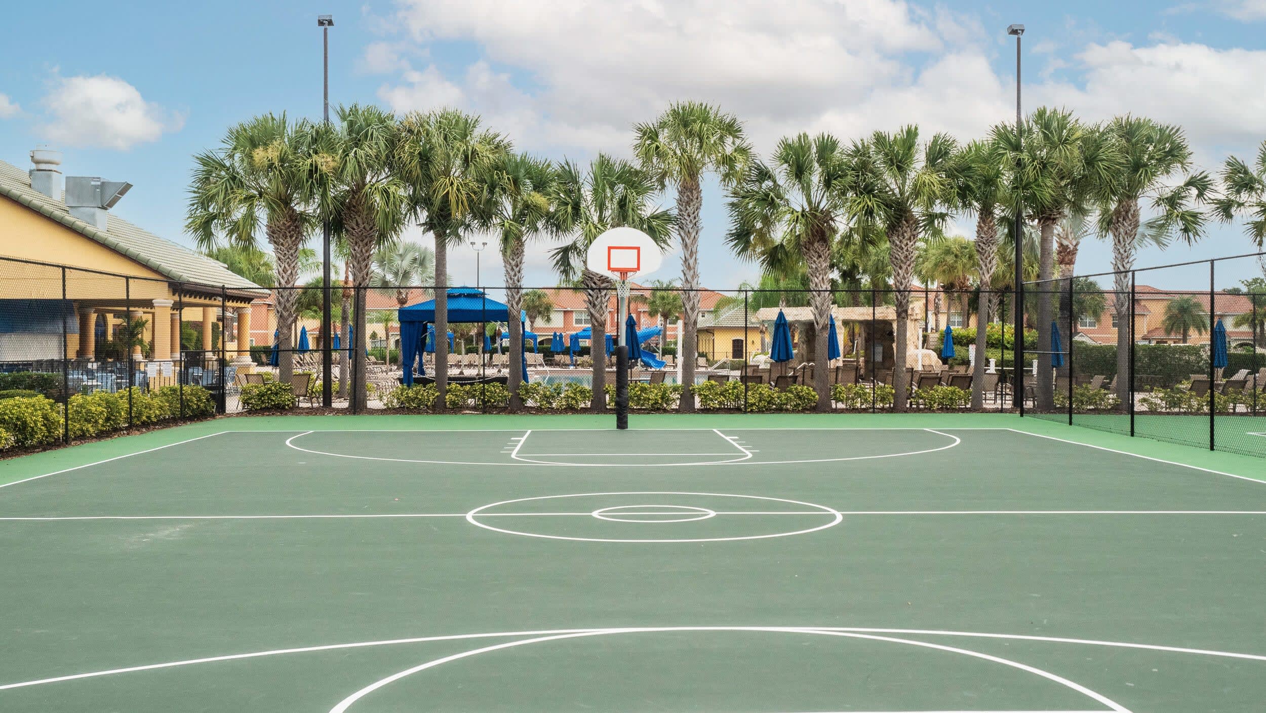 An outdoor basketball court with a green surface and white markings, enclosed by a black fence, with palm trees and resort buildings visible in the background under a bright sky.