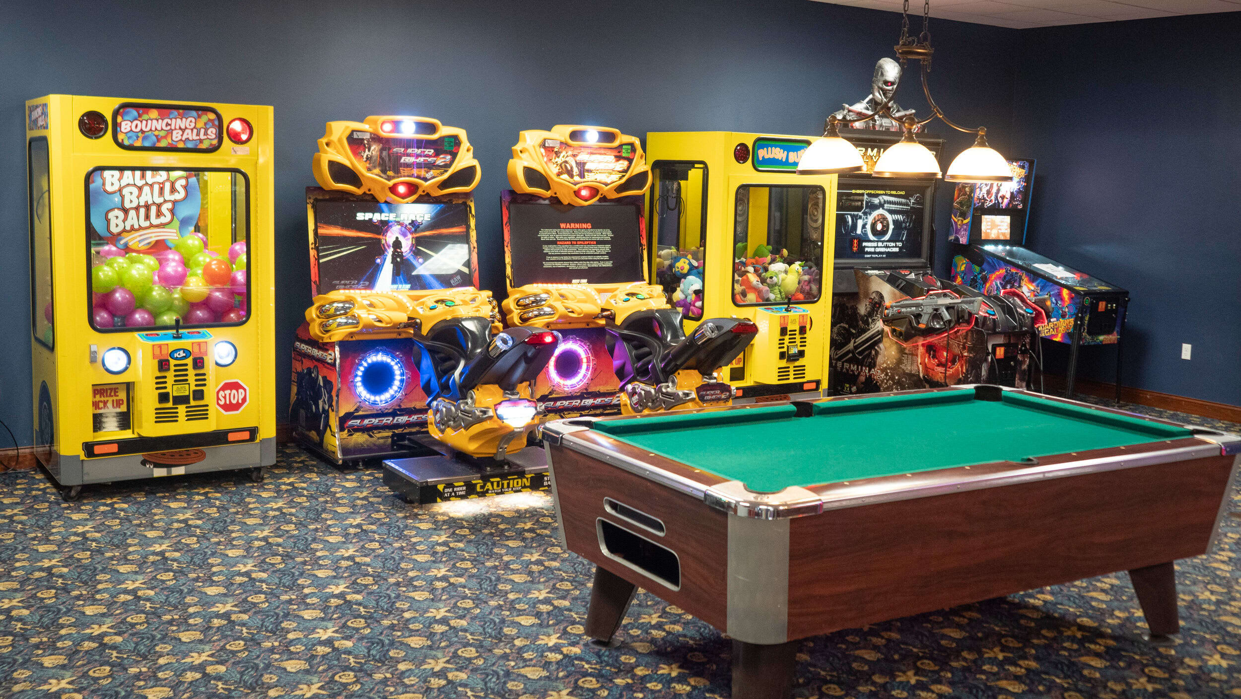 A vibrant game room with dark blue walls and a patterned carpet, featuring a pool table in the foreground and several arcade machines, including "Bouncing Balls" and "Space Invaders", lining the back wall.