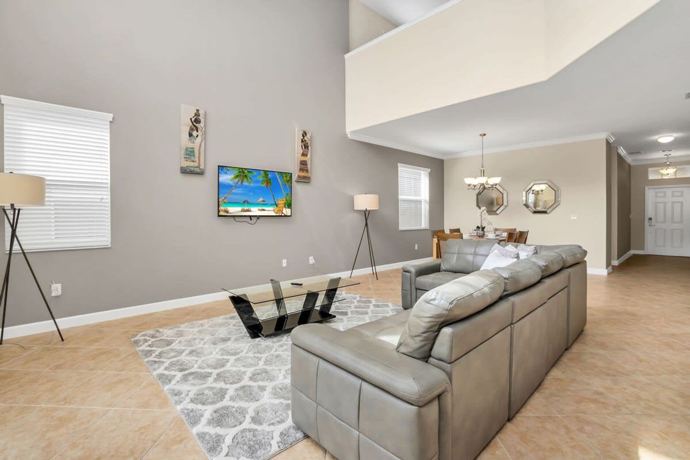 A spacious living room features high ceilings, light gray walls, and a large gray leather sectional sofa facing a wall-mounted TV displaying a beach scene. A glass coffee table sits on a patterned gray and white rug, with two tall floor lamps providing ambient lighting.