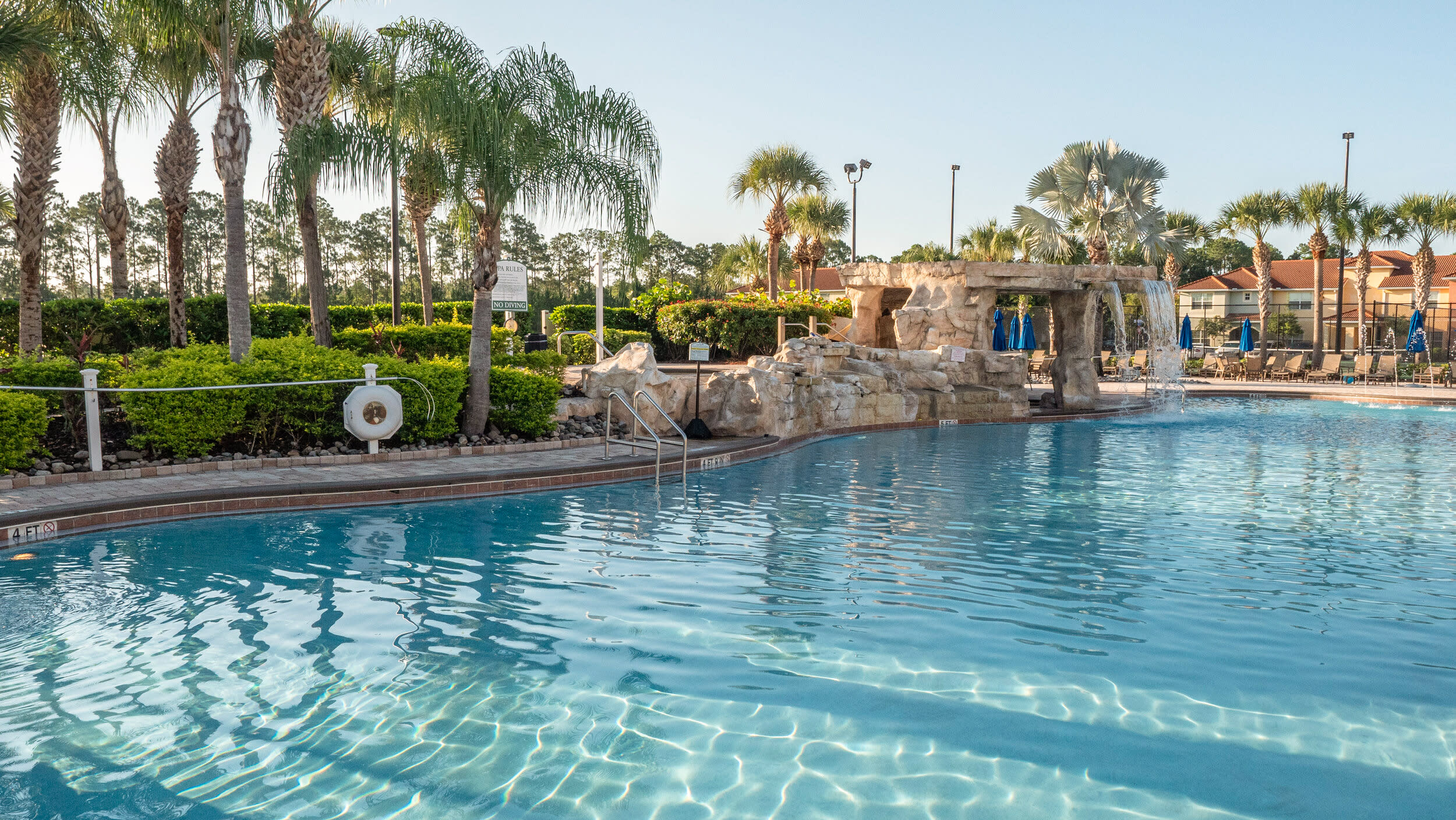 A large communal swimming pool with clear blue water, featuring a rock waterfall structure and surrounded by palm trees and lush landscaping. Lounge chairs are visible in the background, suggesting a resort-style setting.