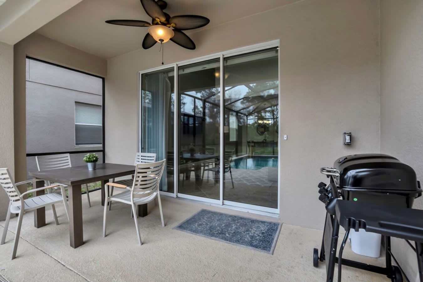 Covered outdoor patio with a dining set and BBQ grill, featuring a ceiling fan and large sliding glass doors leading to the interior, with a view of the screened pool.