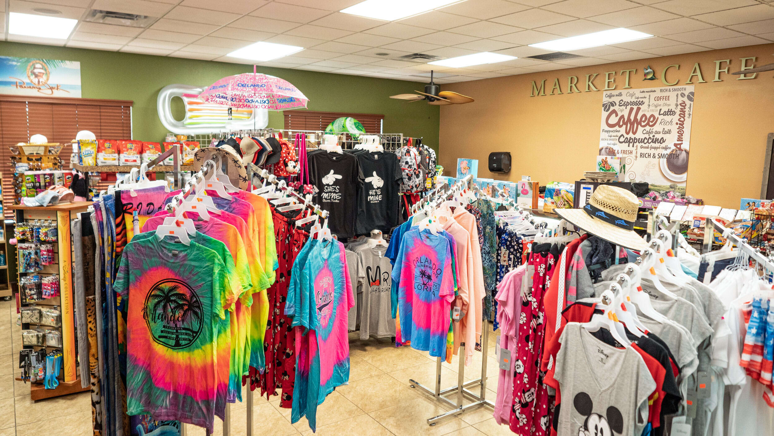 A vibrant indoor market or gift shop filled with racks of colorful t-shirts, hats, and various souvenirs, with a 'Market Cafe' sign on the wall and a bright, welcoming atmosphere.