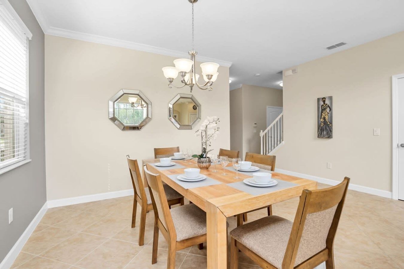 A formal dining area features a light wood rectangular dining table set for six with white place settings, beneath a brushed nickel chandelier. Two octagonal mirrors adorn the light beige wall, and tile floors extend throughout the space.