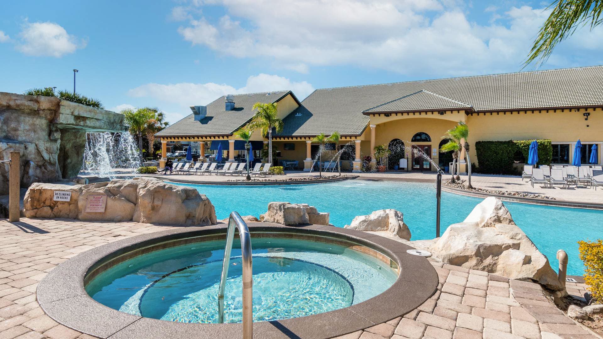 An expansive communal pool area featuring a large swimming pool with a rock waterfall, a circular hot tub in the foreground, and a clubhouse building in the background. The area is surrounded by paved decking and palm trees under a clear sky.