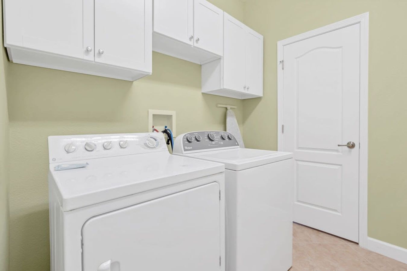 A functional laundry room with light green walls and tiled floors, equipped with a white top-loading washing machine and a matching dryer, both beneath white wall-mounted cabinets for storage. A white door is visible on the right.