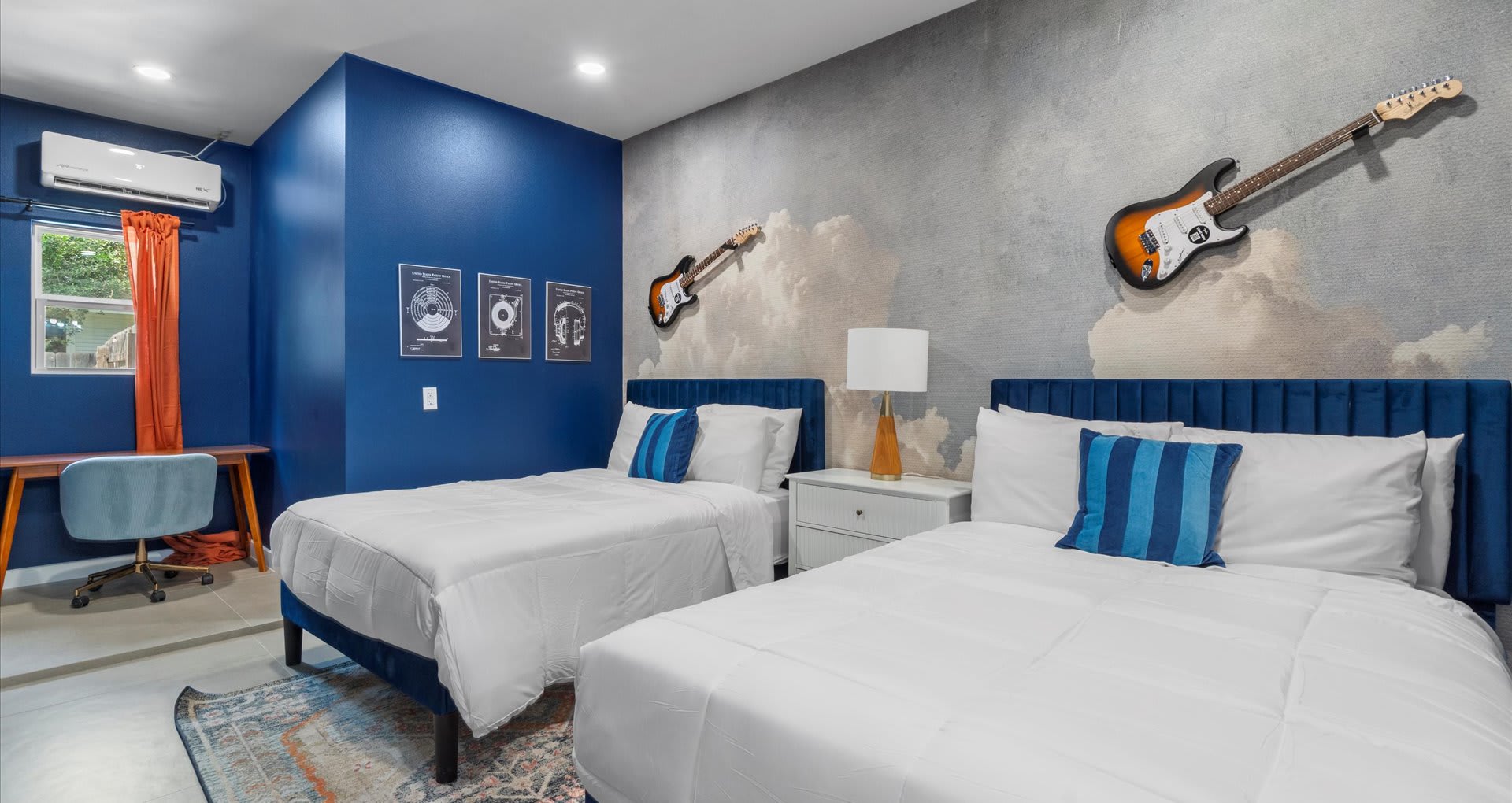 Bedroom with two twin beds featuring blue velvet headboards and white bedding, set against a gray cloud-patterned accent wall with two electric guitars mounted as decor. The room has a bold blue accent wall and a small desk area with a light blue chair.